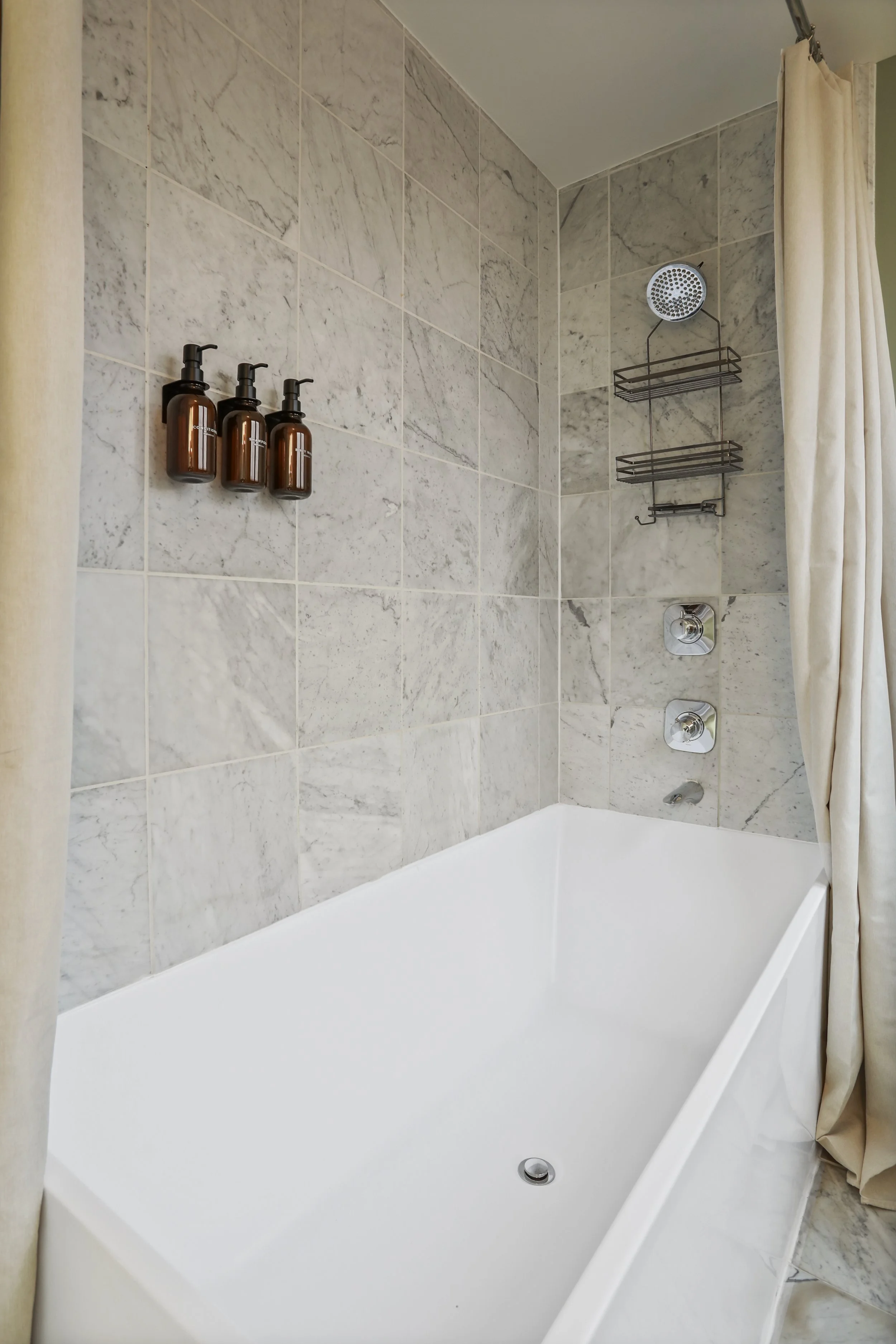 Bathroom with a bathtub, tiled walls, and a showerhead with shelves. Cream-colored curtain on the right.