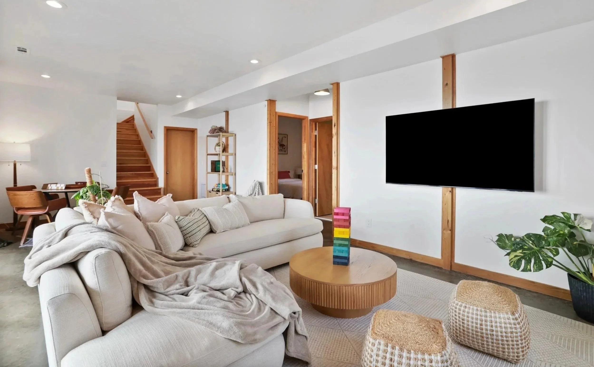 Living room with a white sofa, a round wooden coffee table with a stack of colorful blocks, a flat-screen TV mounted on a wall with wooden trim, a large potted plant, and a corner with a bookshelf and a staircase in the background.