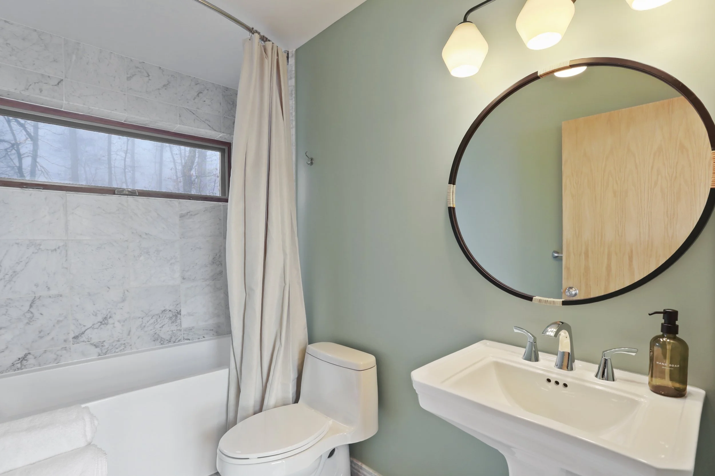 A modern bathroom with a porcelain vanity and faucet, a round mirror, a white toilet, a window with a marble-like tile border, a shower with a curtain, and a bottle of hand soap on the sink.