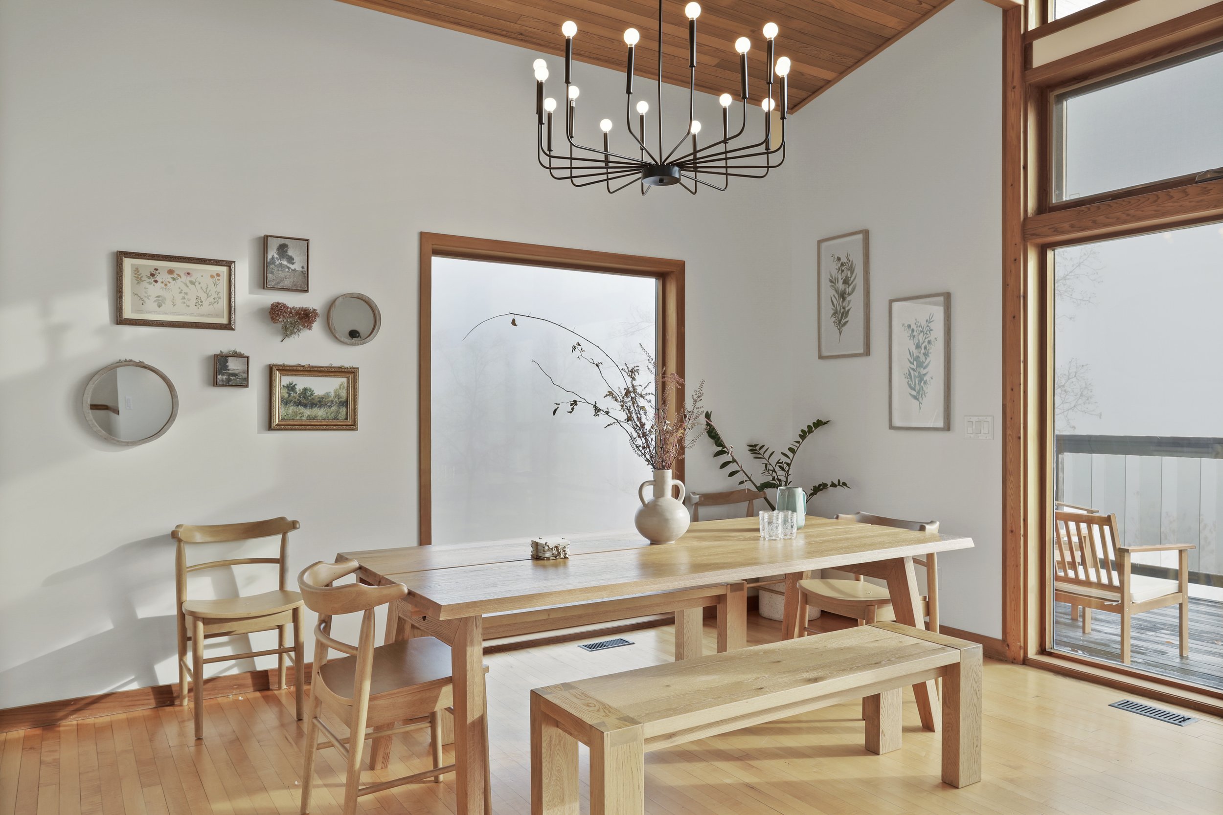 Dining room with wooden table, benches, and chairs, decorated with vases and plants, featuring a modern chandelier, white walls, framed botanical art, and a large window with a sliding door leading to a balcony.
