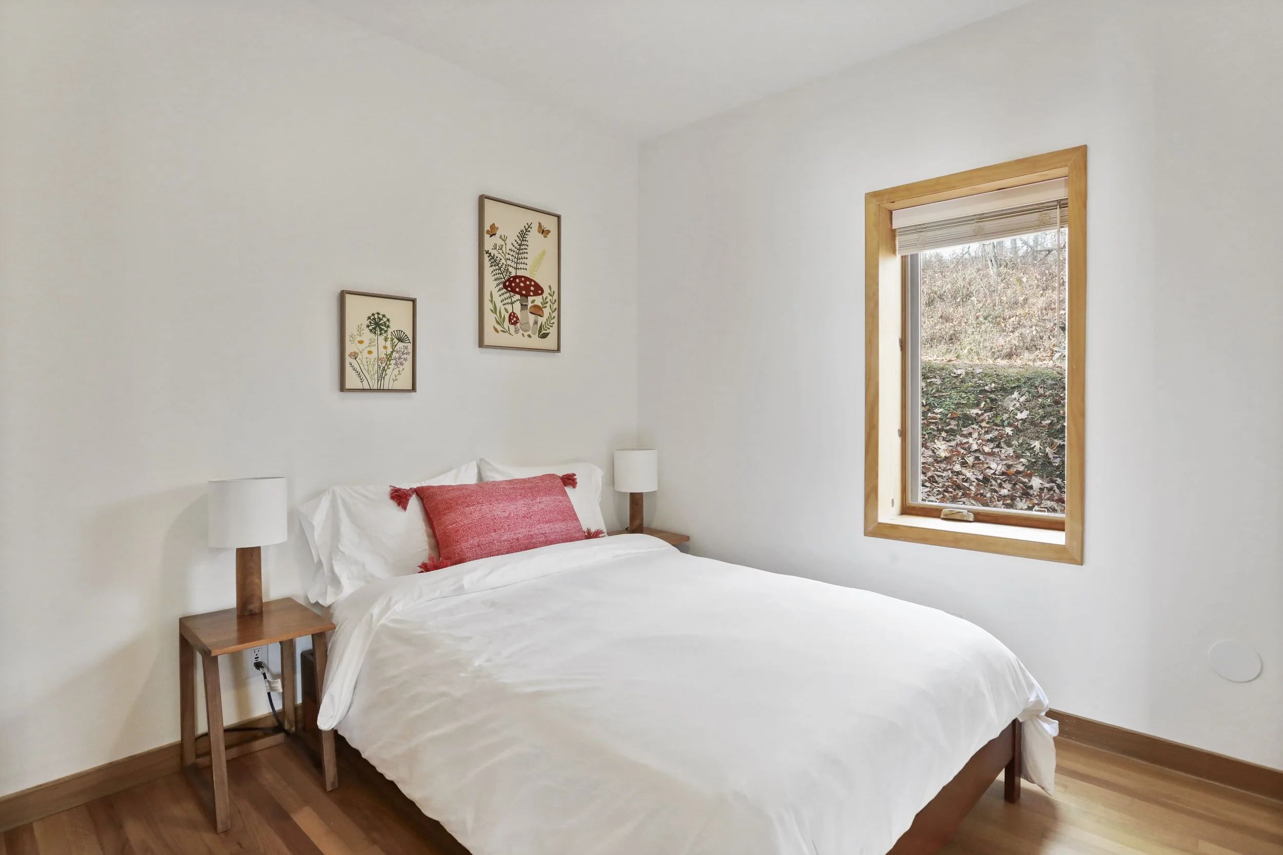 A bedroom with white walls, a bed with white bedding and a red pillow, two wooden nightstands with white lamps, and a window with a view of fallen leaves outside.