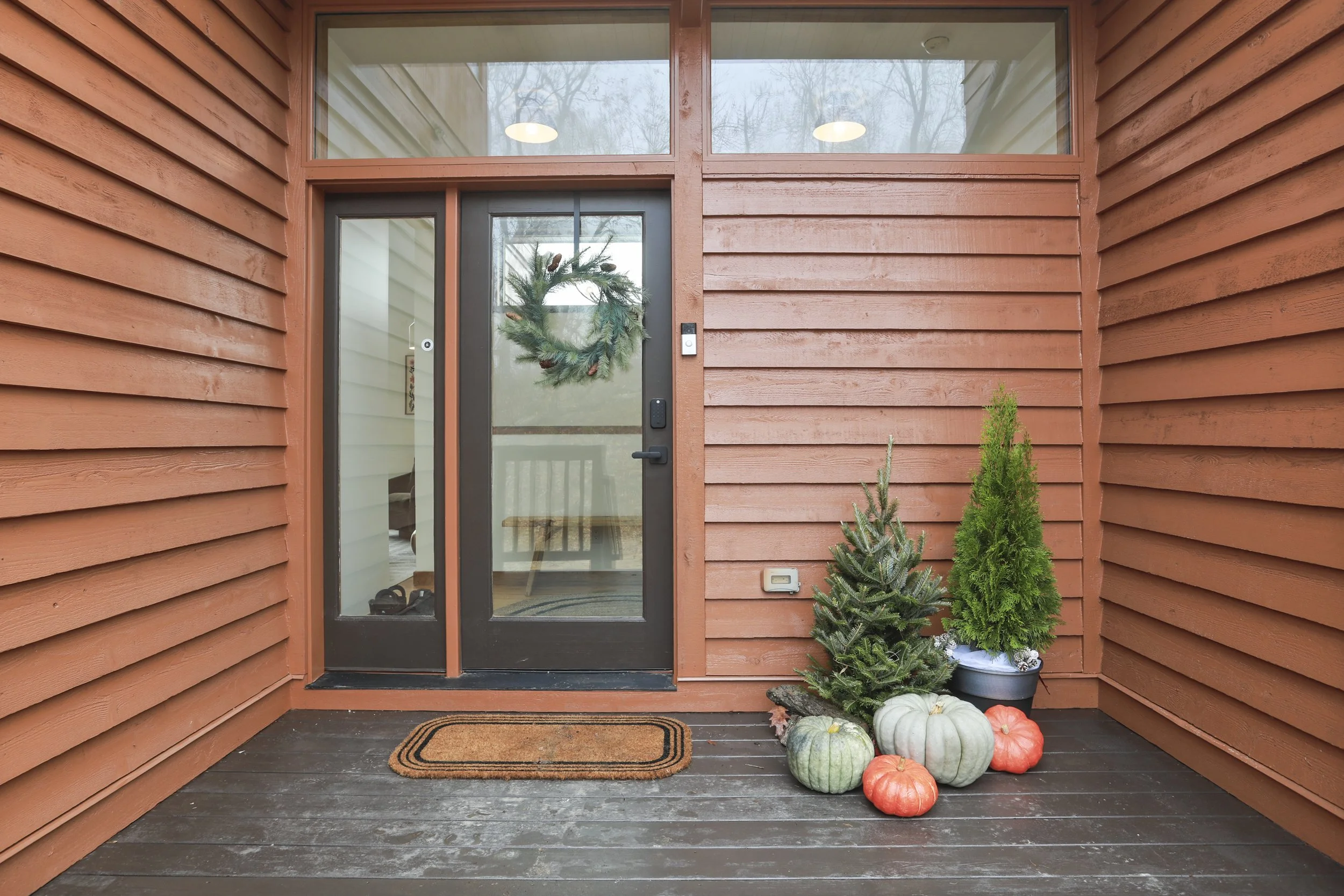 Front porch with orange wood siding, a glass door with a holiday wreath, and fall pumpkins and potted mini trees outside.