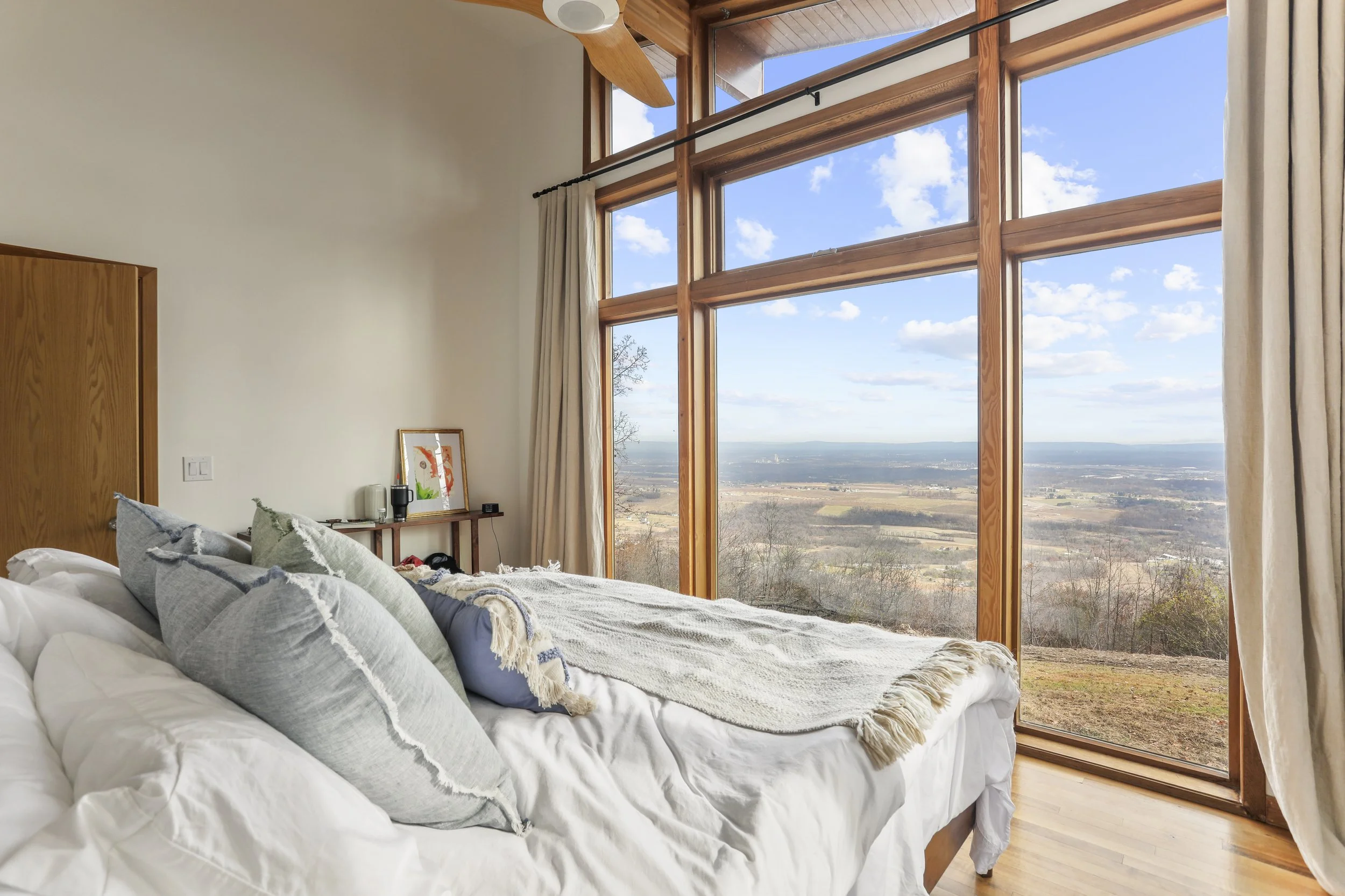 A bedroom with a large floor-to-ceiling window showing a scenic view of the countryside, with blue sky and clouds, wooden framing, and curtains on the sides.