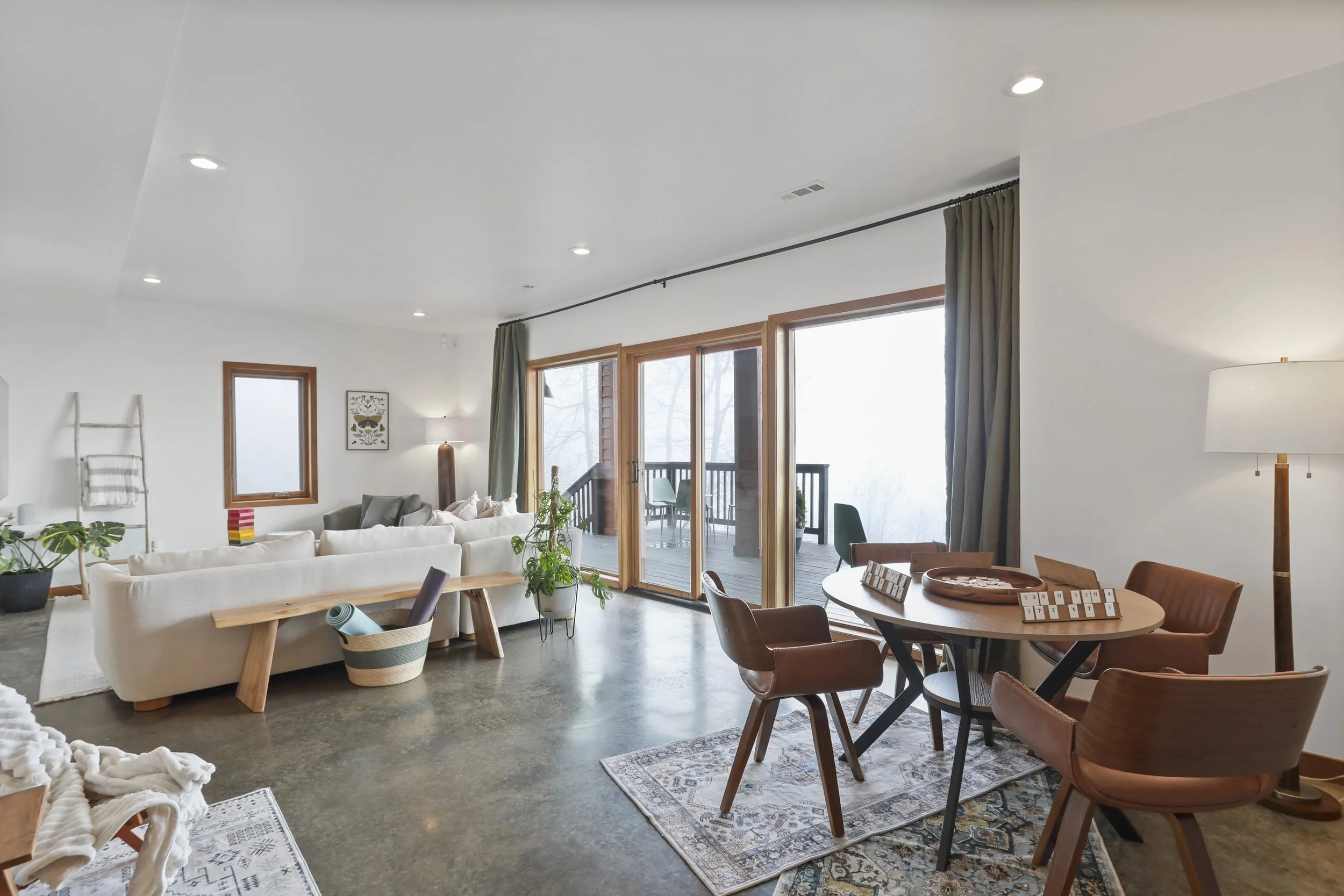 A spacious living and dining area with large sliding glass doors leading to a balcony. The room features a white sofa, a wooden dining table with four chairs, several plants, and decorative rugs. Recessed lighting and a tall floor lamp brighten the s