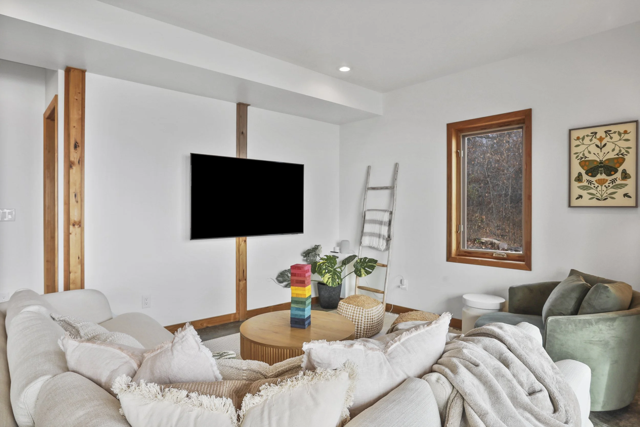Cozy living room with white walls, a beige sectional sofa with white pillows, a round wooden coffee table with colorful Jenga blocks, a green armchair, a large window with natural light, a wall hanging with butterfly and floral design, a ladder decor