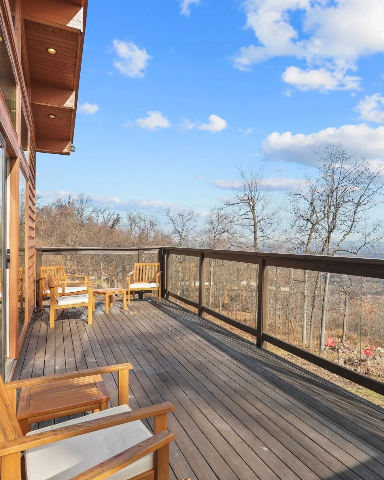 A spacious outdoor wooden balcony with several wooden chairs with white cushions, surrounded by a glass railing overlooking a landscape of leafless trees under a blue sky with scattered clouds.