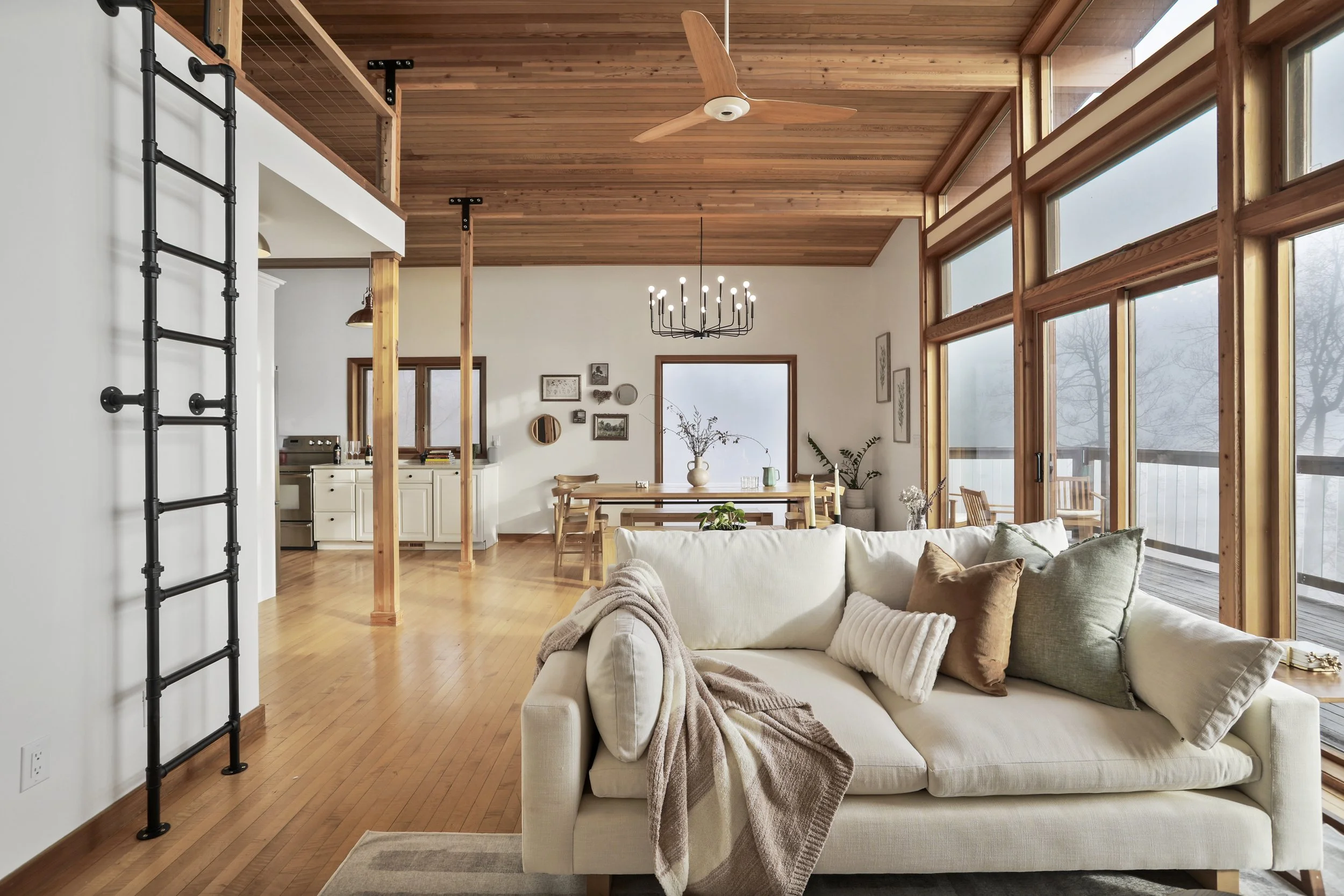 Bright living room with large windows, wooden ceilings, and a white sofa with multiple pillows. Dining area with a wooden table and chairs, decorative wall art, and a chandelier. Some plants and outdoor balcony visible.