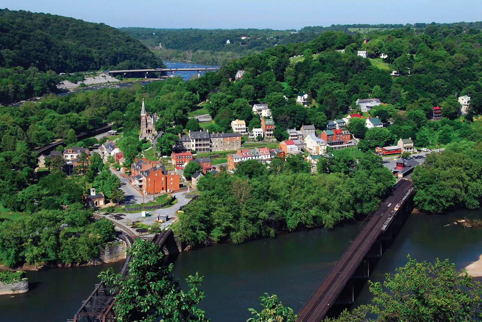 A scenic aerial view of a small town situated among green hills with a river running through it, featuring a bridge and a train crossing.
