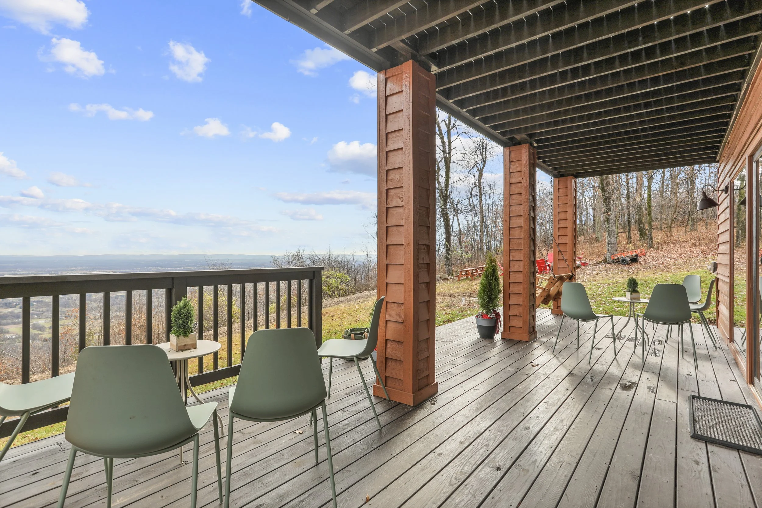 Empty outdoor wooden deck with chairs and tables, overlooking a scenic landscape with trees and a cloudy sky.