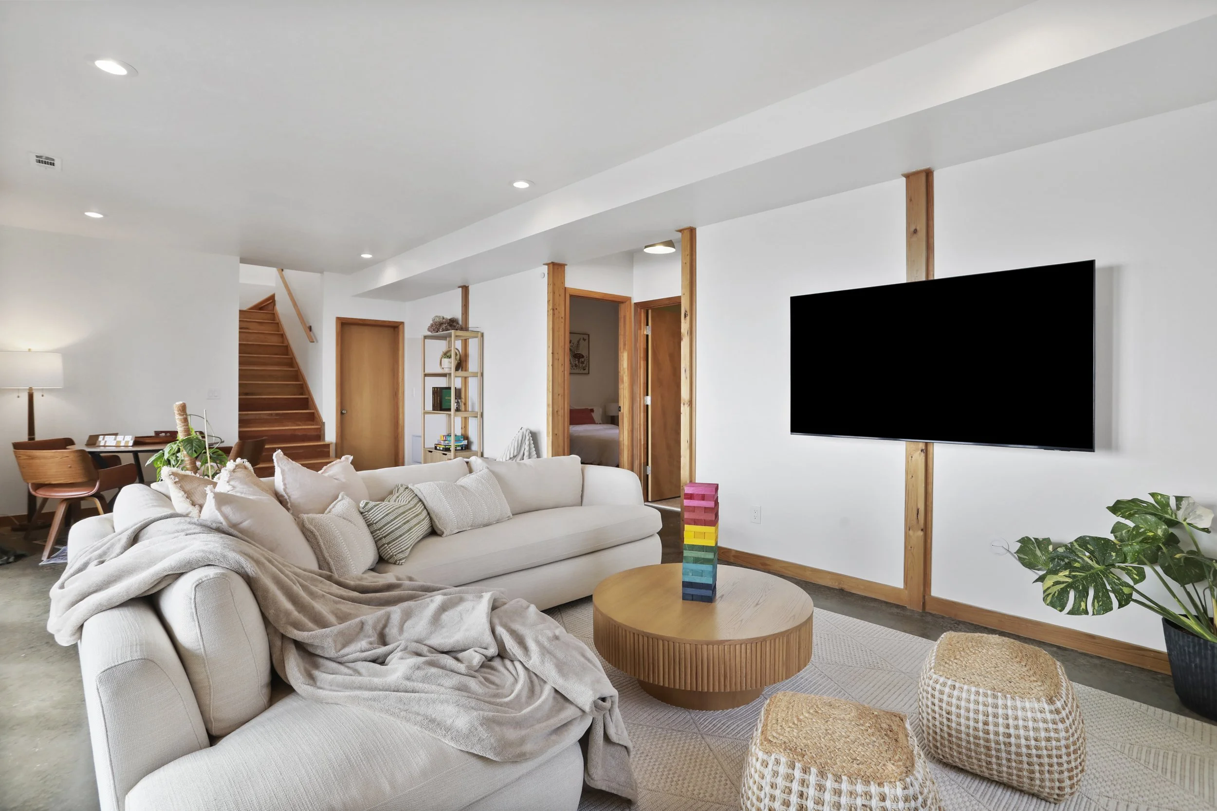 Modern living room with white sofa, beige blanket, stacked colorful game pieces on round wooden coffee table, potted plants, wall-mounted TV, and wooden trim accents.