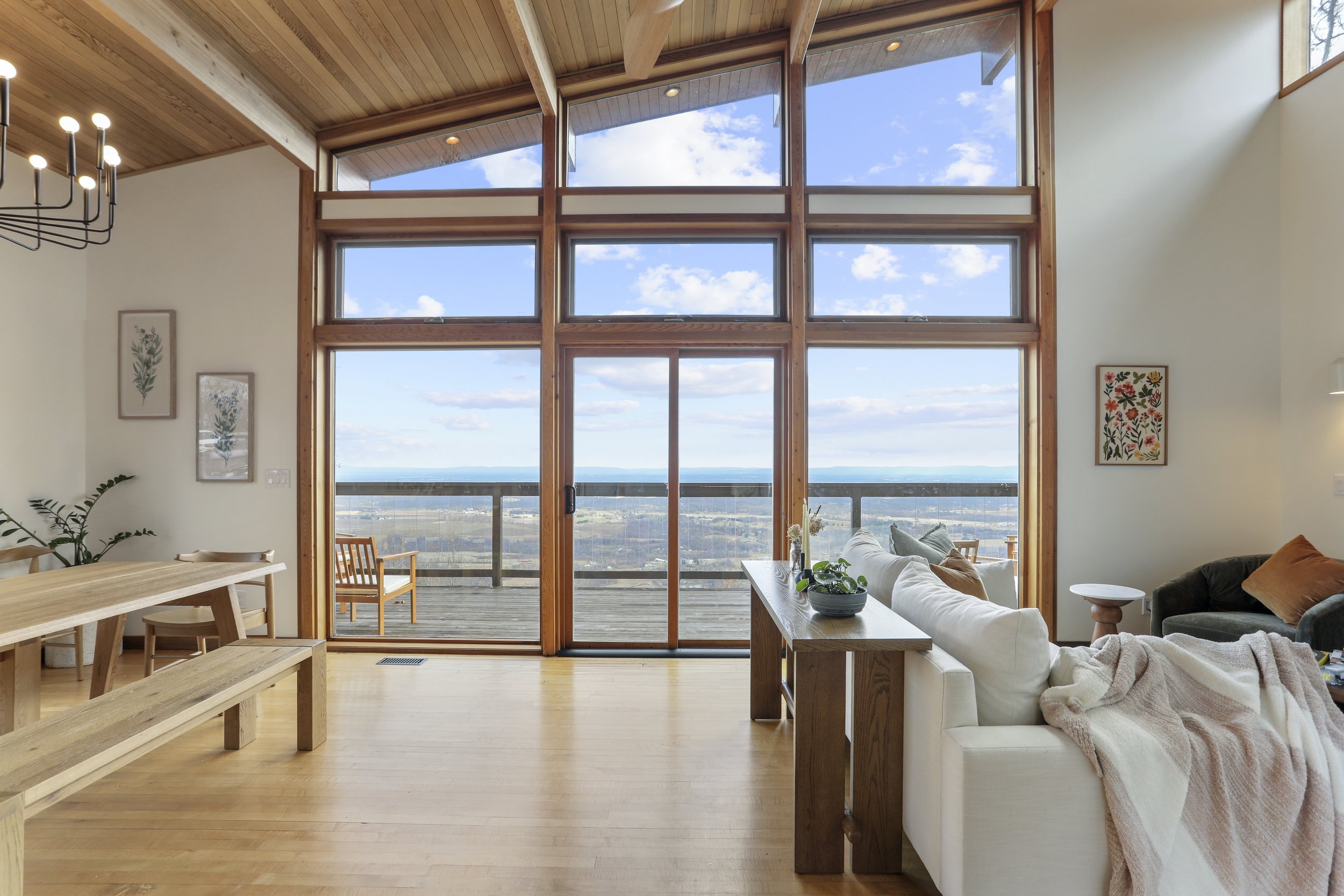 Living room with large windows and sliding glass door overlooking a balcony and scenic landscape, wooden beams, modern chandelier, beige sofa with throw blanket, armchair, and artwork on the walls.