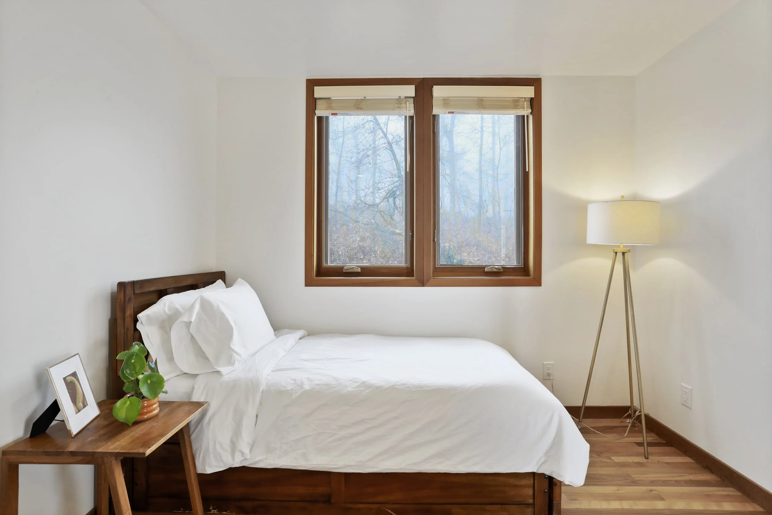A bedroom with a neatly made bed, a side table with a framed picture and plant, large windows with a view of trees, and a standing lamp.