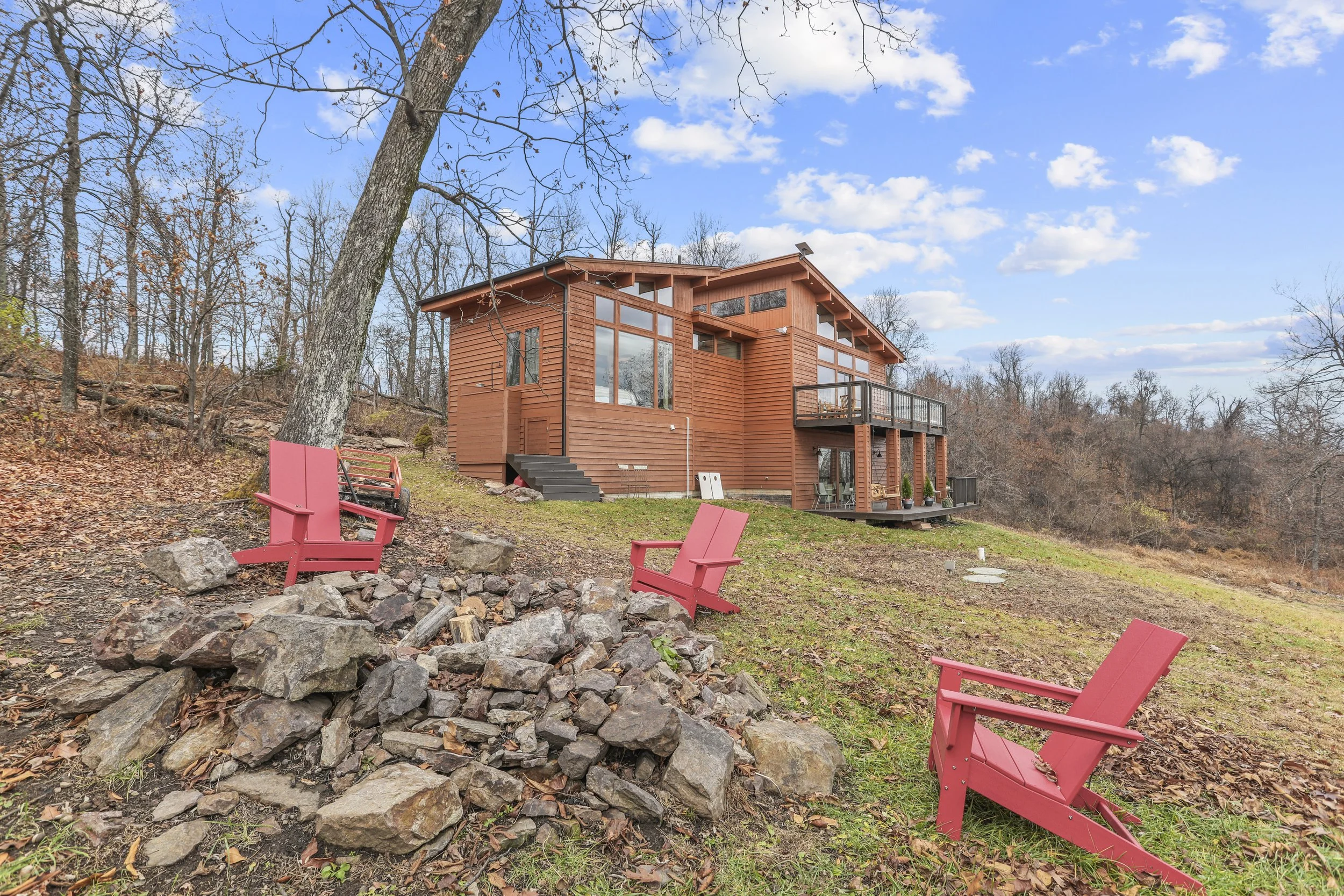 A wooden house on a sloped yard with three pink Adirondack chairs around a small fire pit and leafless trees in the background under a partly cloudy sky.