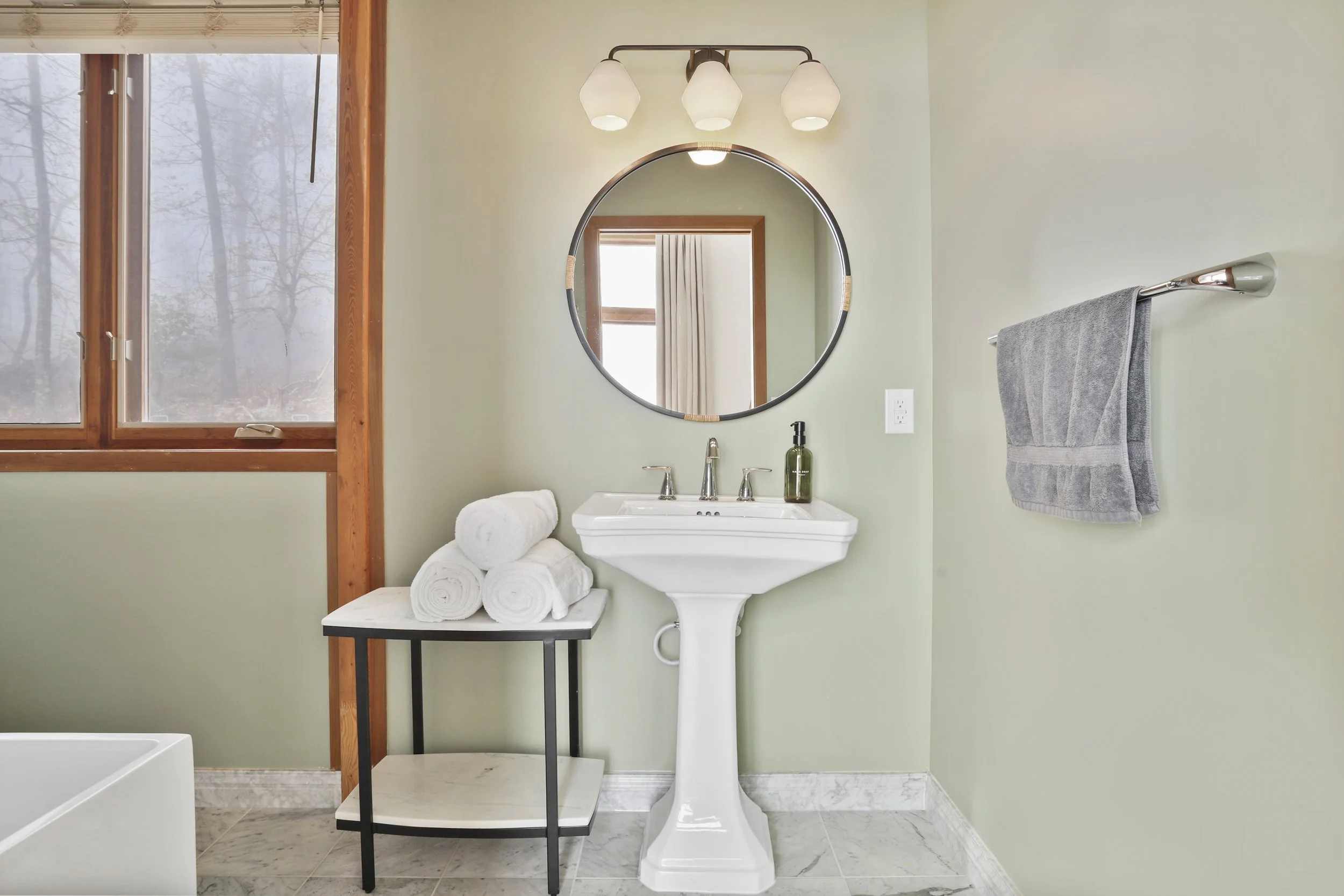 Bathroom with a pedestal sink, round mirror, towel rack with gray towel, window, and a small table with rolled white towels.