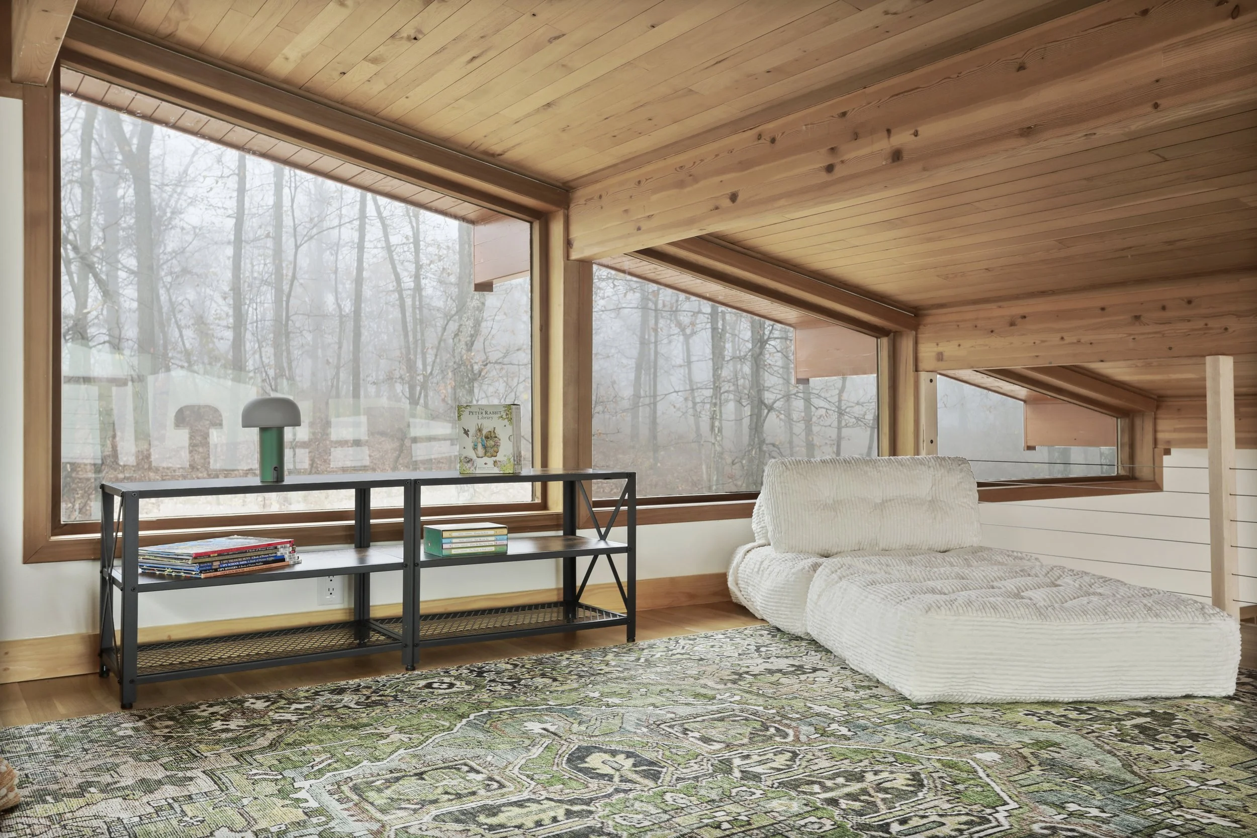 Room with large sloped glass windows revealing a wooded outdoor scene, a modern black metal bookshelf, a white floor sofa, patterned area rug, and wood-paneled ceiling and walls.