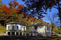 Two houses with mature trees in front, one white with arches and the other with a sloped roof, on a sunny day with a clear sky.