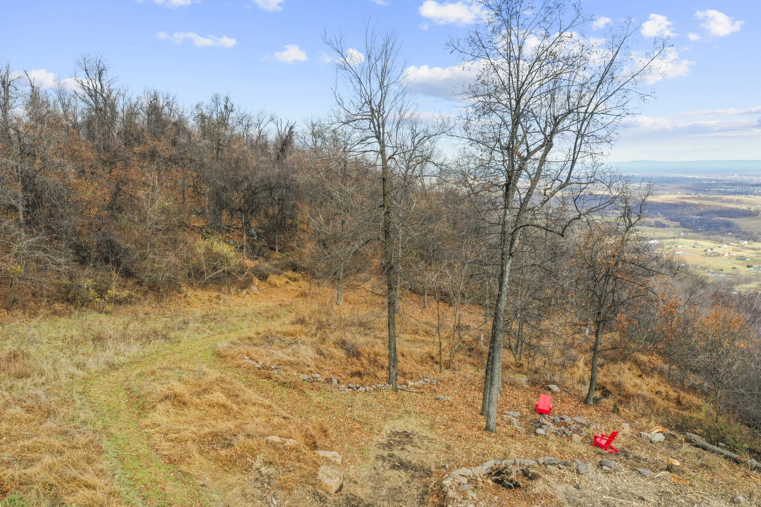 A scenic autumn landscape with a dirt path, leafless trees, and autumn foliage on the hillside. Two red Adirondack chairs are situated near rocks at the edge of the slope, overlooking fields and a distant valley under a partly cloudy sky.