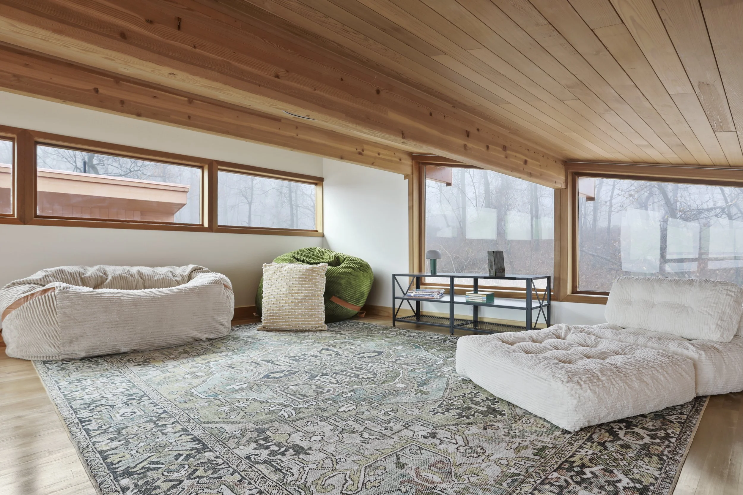Modern living room with large windows, wooden ceiling, beige couches, green bean bag chair, patterned rug, and a black metal table with books and decor.