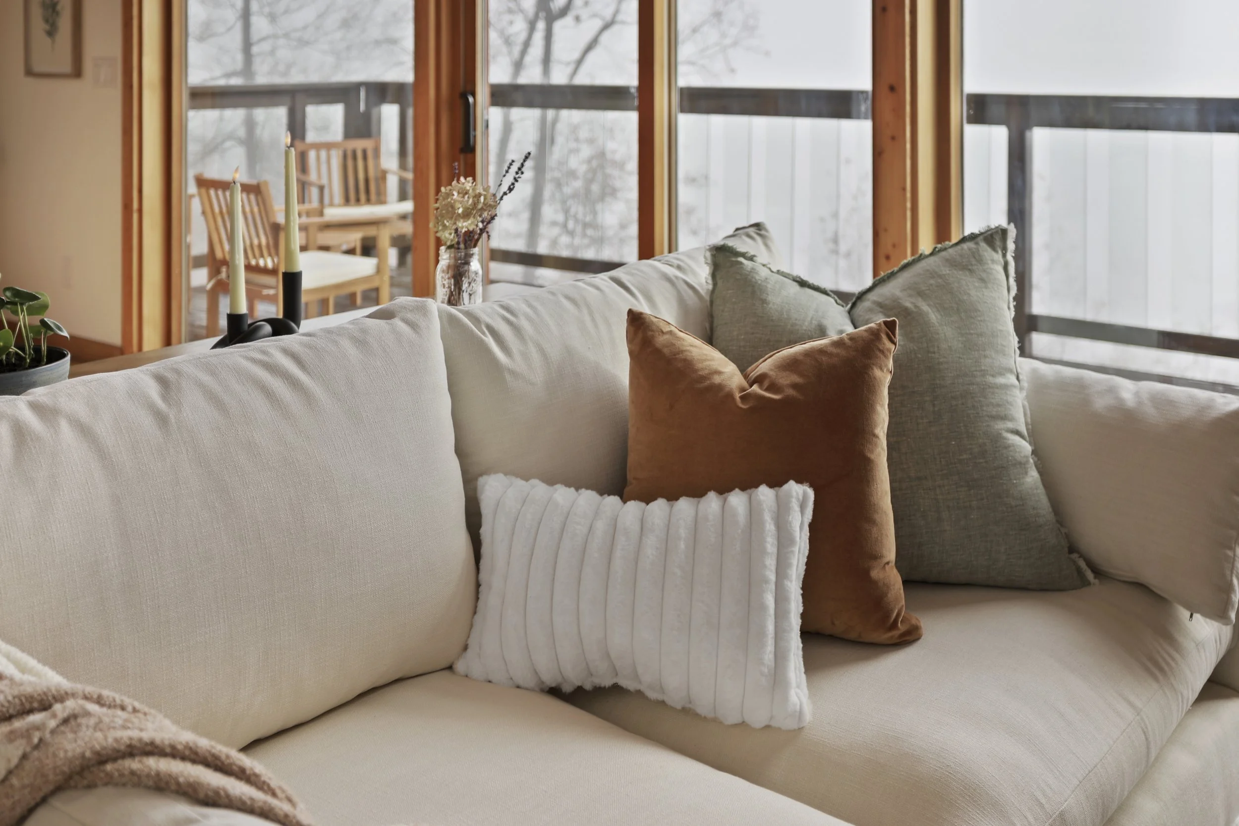 A beige sofa with four pillows: a white, ribbed pillow, a tan velvet pillow, and two grey pillows. Behind the sofa, large windows reveal a snowy outdoor scene with trees, a balcony, and a wooden dining table with chairs.