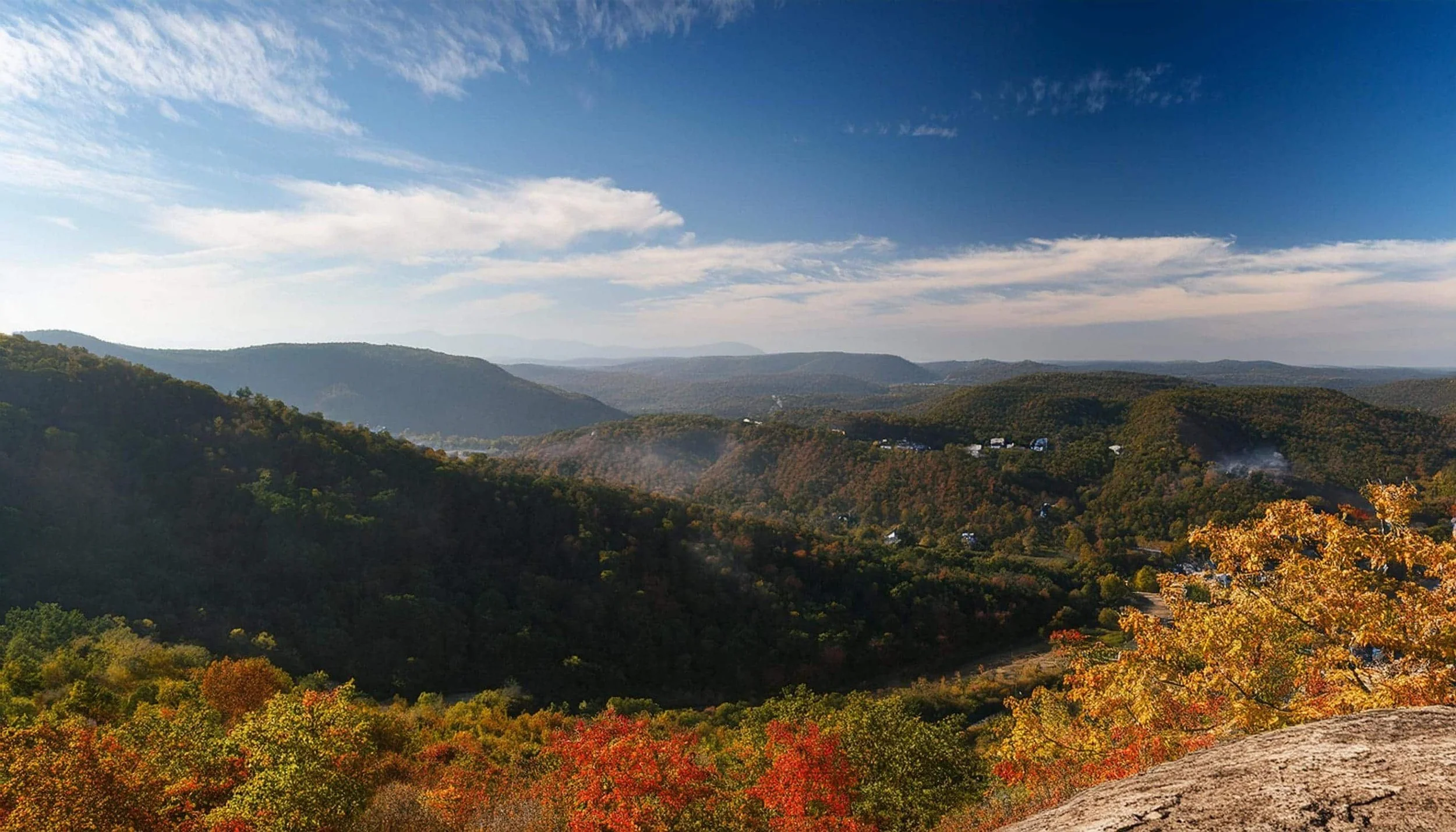Scenic view of rolling mountains with a partly cloudy sky, autumn foliage, and some smoke rising in the distance.