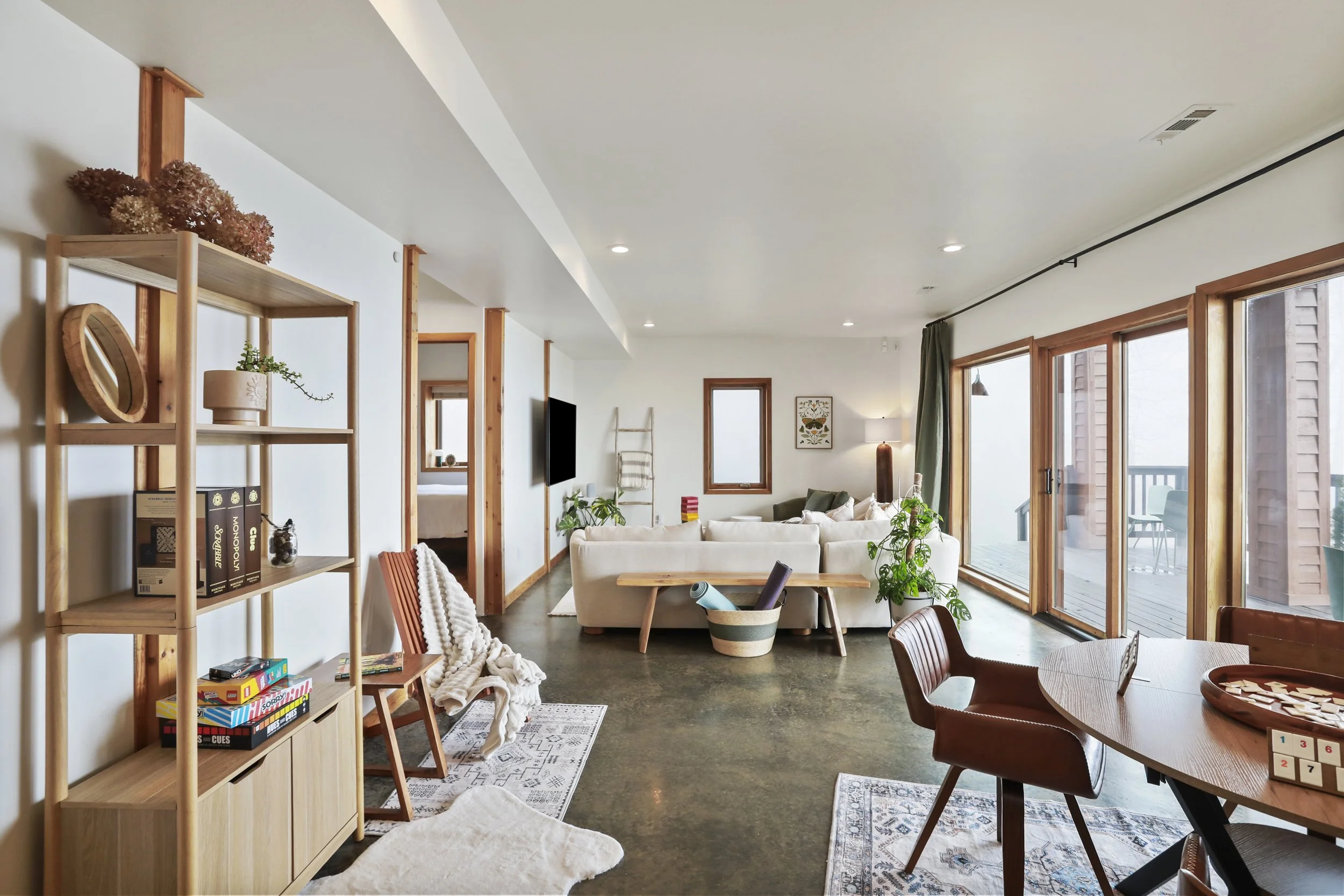 Open living room with white sofa, wooden shelves, game board, and plants, leading to a balcony with sliding glass doors and outdoor furniture.