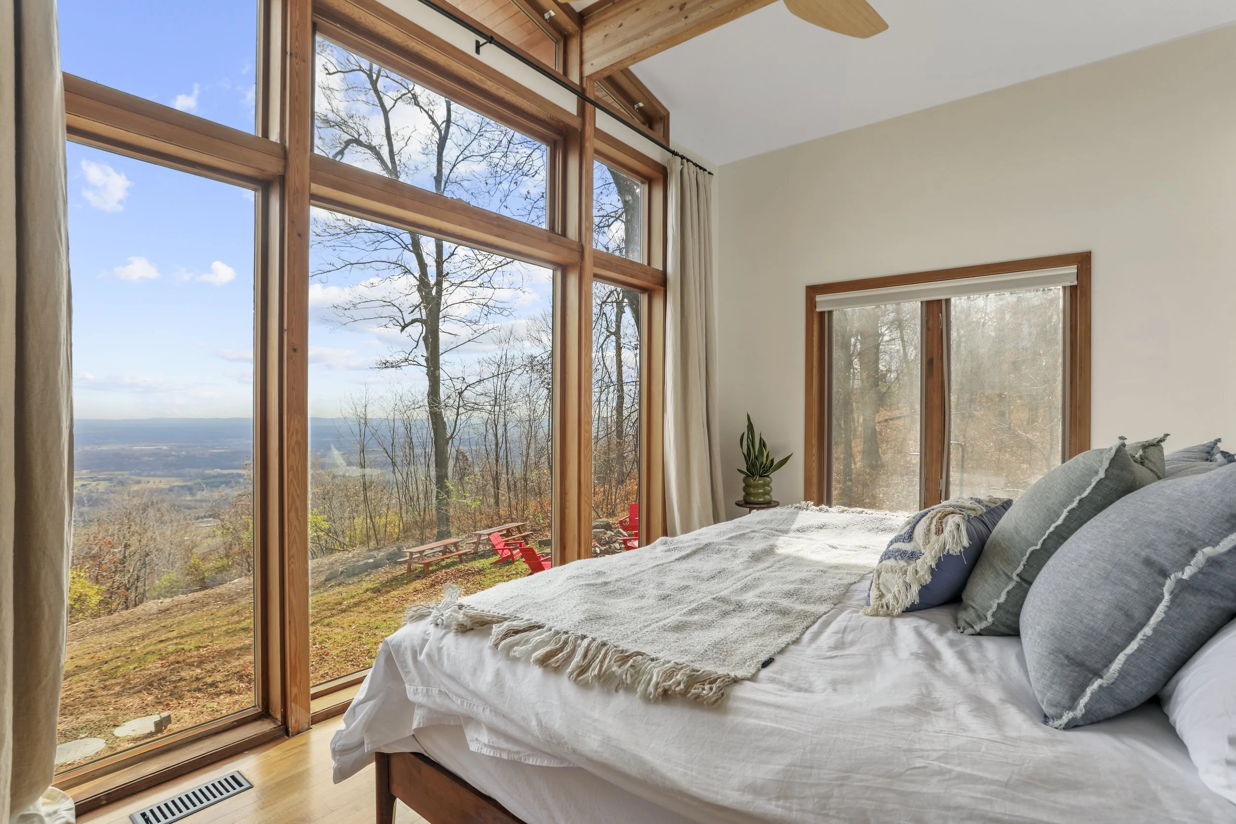 Bedroom with large glass windows overlooking a scenic landscape with trees and a view of the horizon, bed with pillows and blanket, potted plant on bedside table.