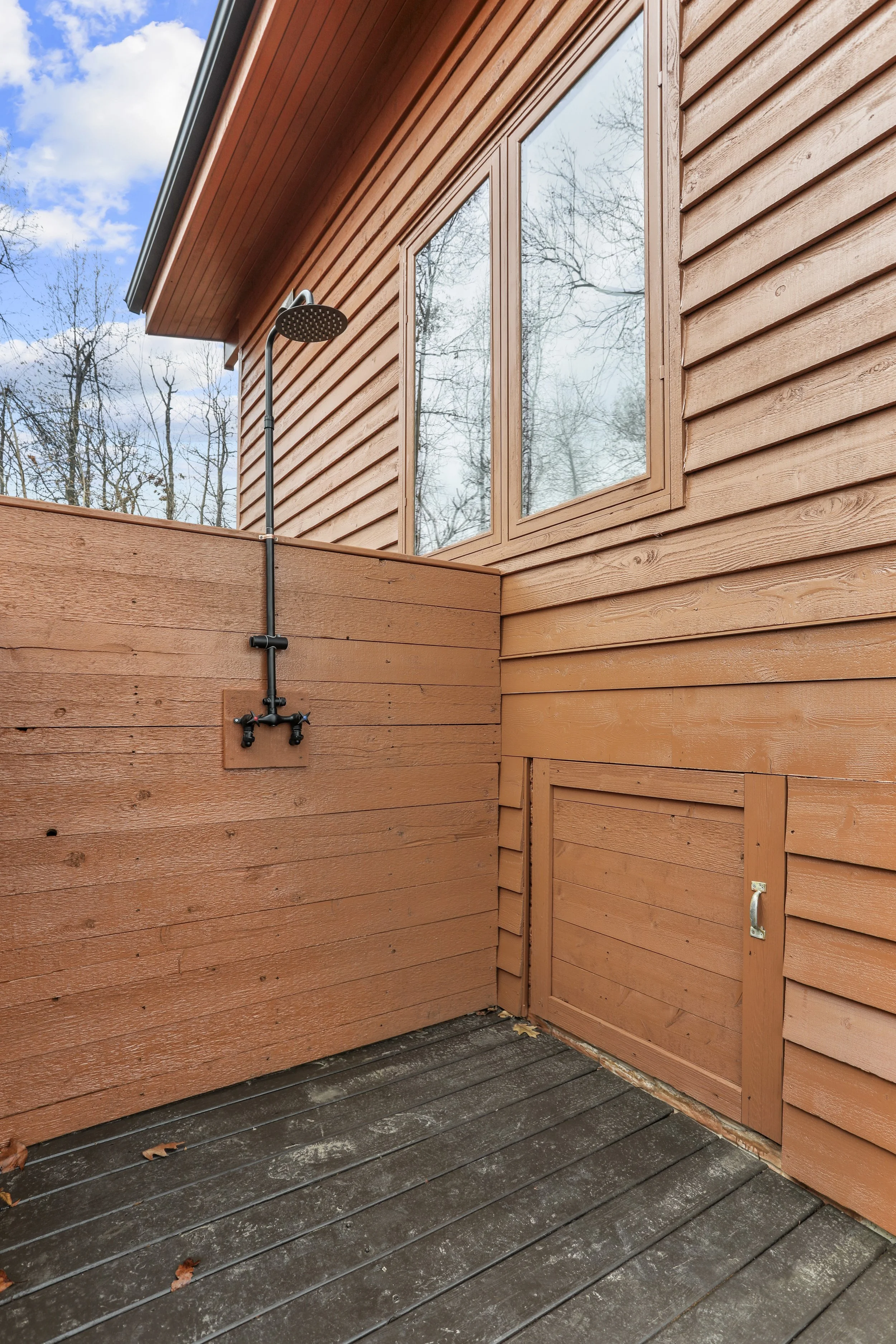 Outdoor shower with black showerhead and pipes on a brown wooden wall on a deck with large windows and a view of leafless trees against a cloudy sky.
