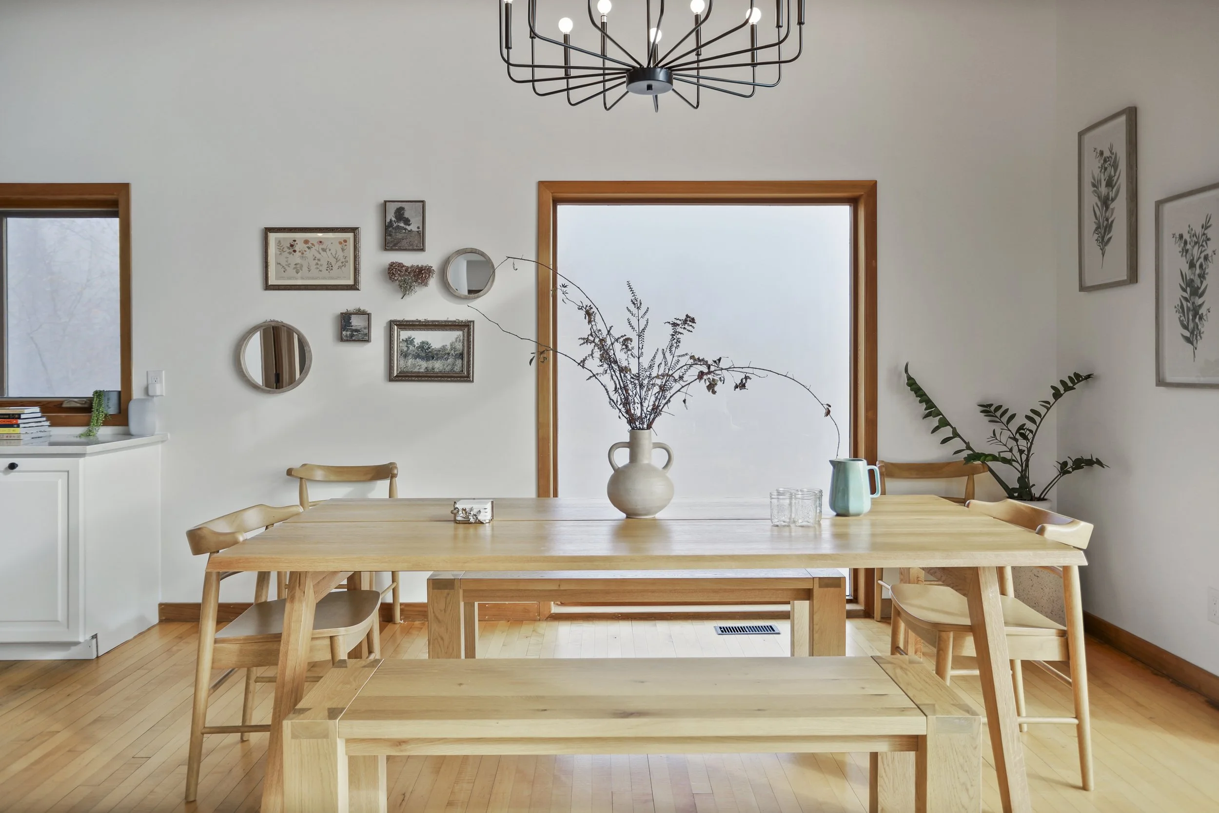 A dining room with a wooden table, benches, and chairs, decorated with a large vase of dried branches in the center, a smaller jug, and glasses. The room has white walls, framed artwork, a metal chandelier, and natural light from a large window and sliding glass door.