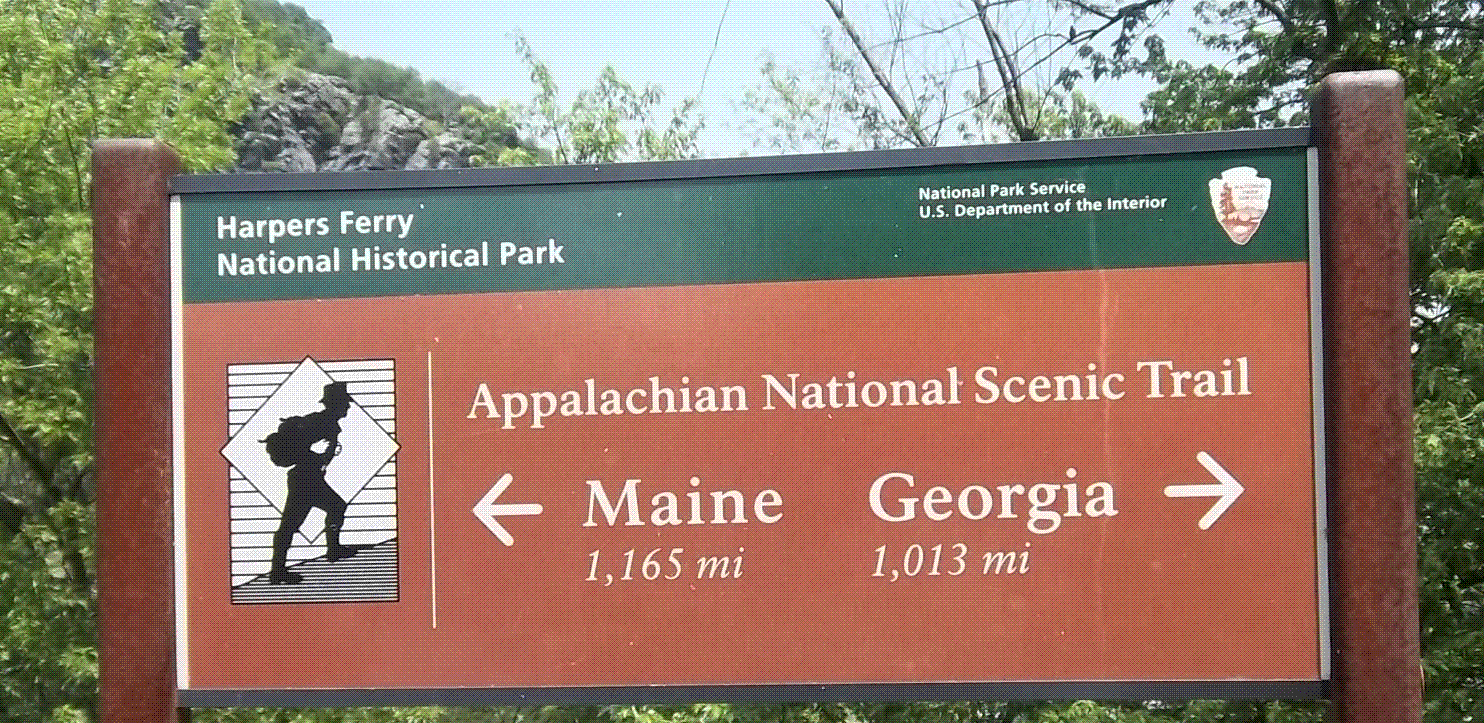 Sign for the Appalachian National Scenic Trail at Harpers Ferry, indicating directions to Maine and Georgia, with distances of 1,165 miles to Maine and 1,013 miles to Georgia, featuring a hiker icon and the logos of the National Park Service and U.S. Department of the Interior.