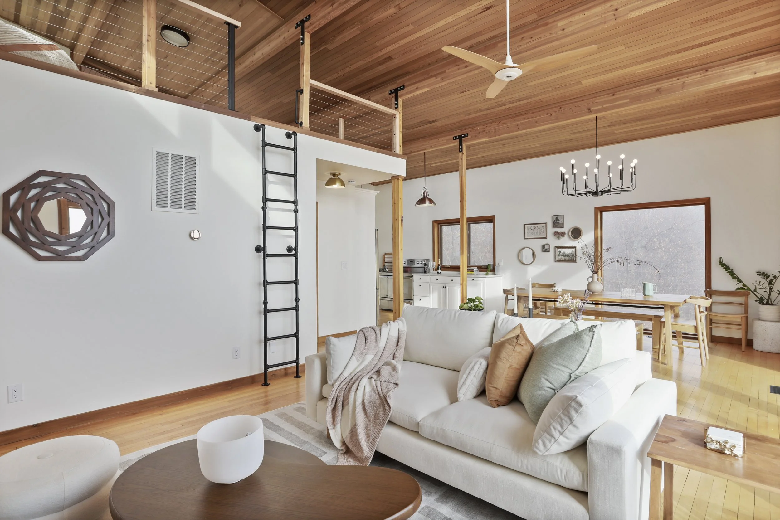 Open-concept living room with white sofa, wooden ceiling, and kitchen in the background; loft area with railing and ladder, natural light, and minimalist decor.