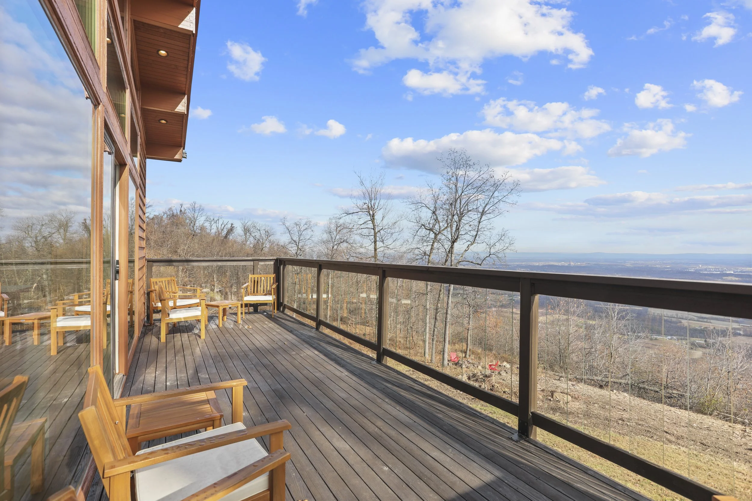 Wooden balcony with several chairs and a glass railing, overlooking a scenic landscape with trees and a distant view of the horizon under a partly cloudy sky.