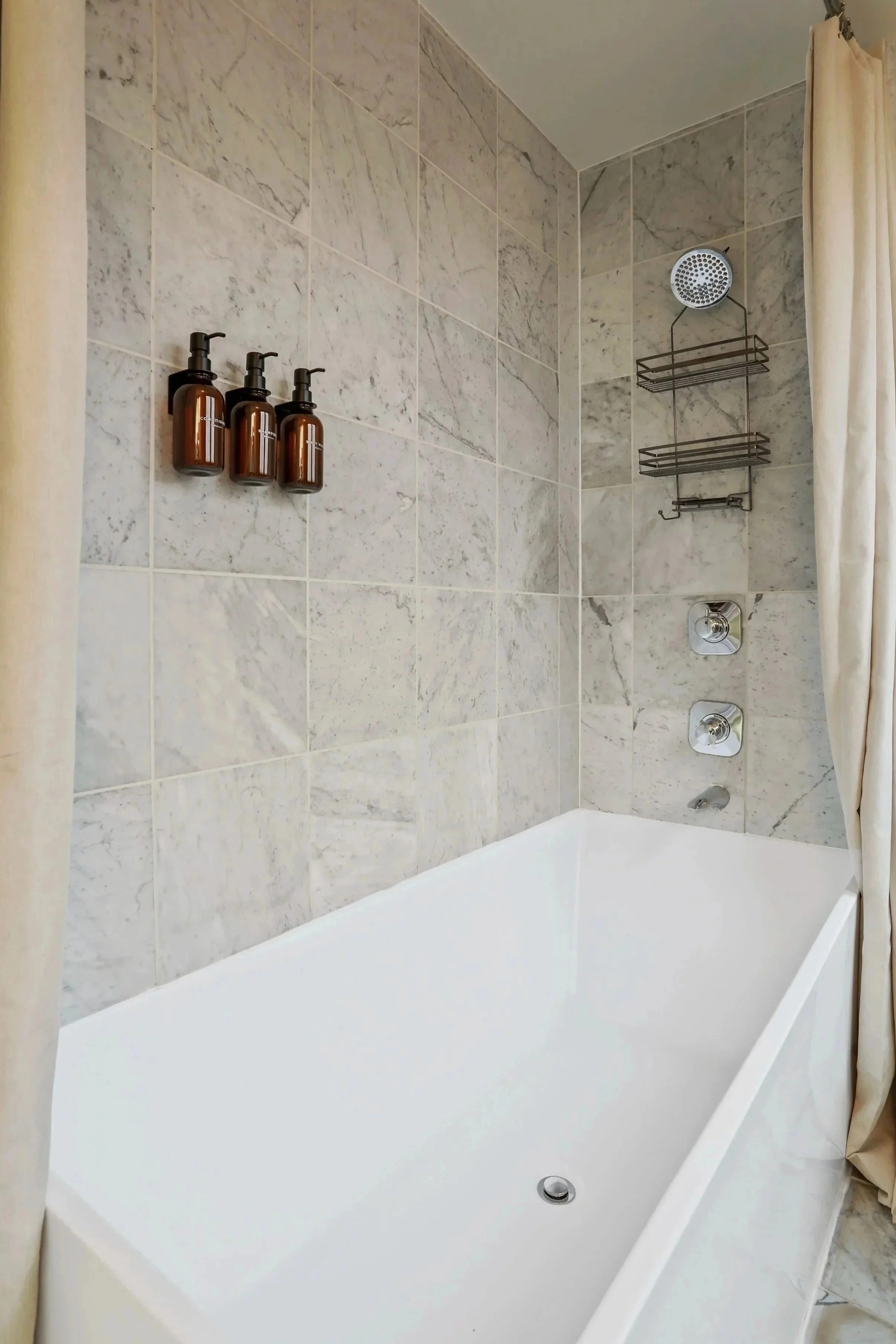 Bathroom with a white bathtub, marble tiled walls, brown soap dispensers, metal shower rack, showerhead, control knobs, and curtain.