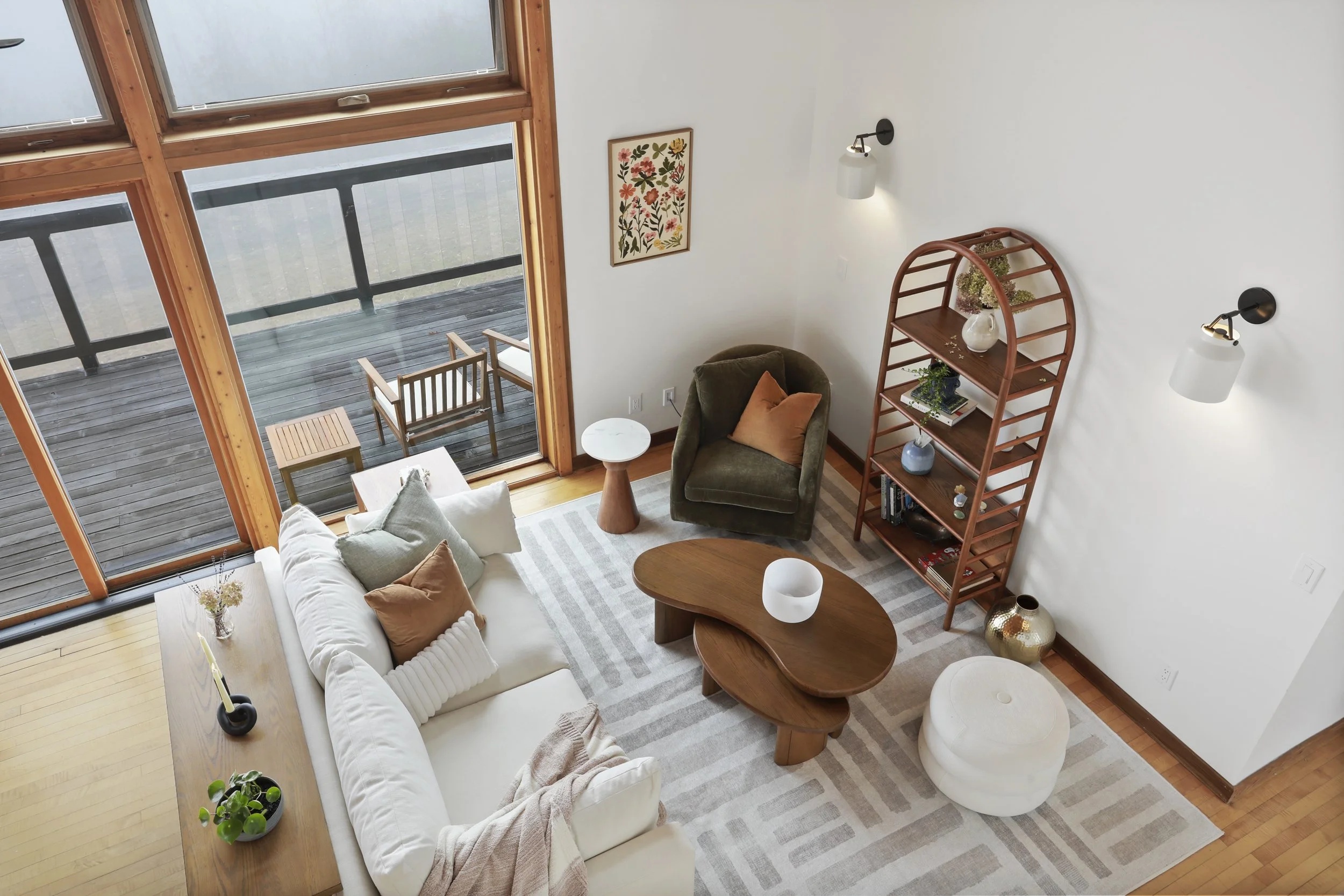 Living room with large windows leading to a balcony, white sofa with pillows, a green armchair, a wooden coffee table, a tall wooden bookshelf with decorative items, two wall-mounted lamps, a small side table, and a striped area rug.