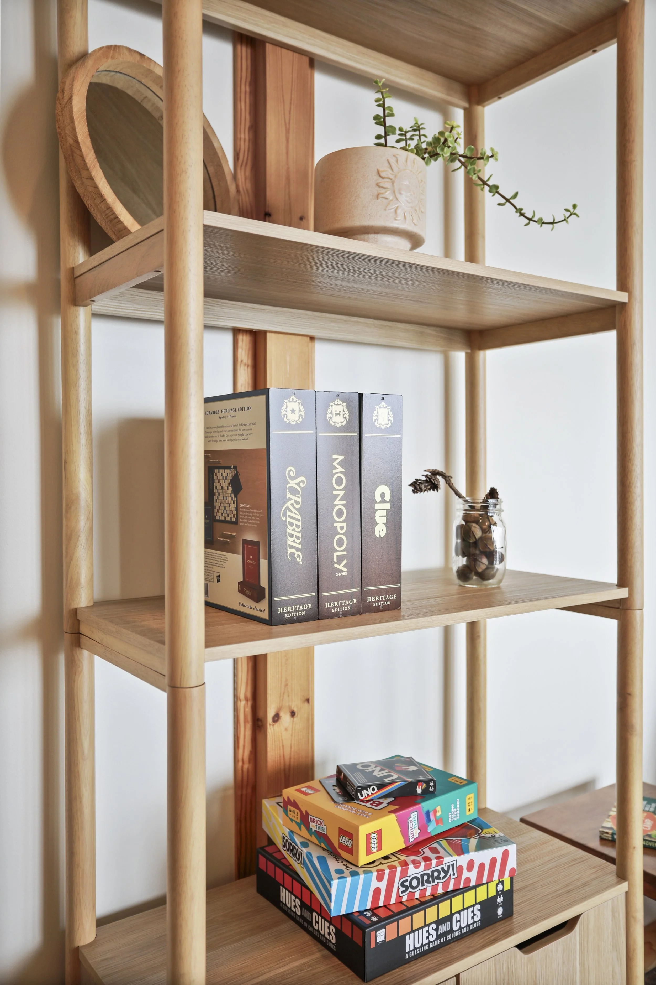 Wooden shelving unit with board games, books, a jar with a sprig, and a decorative wooden dish with a sun design.