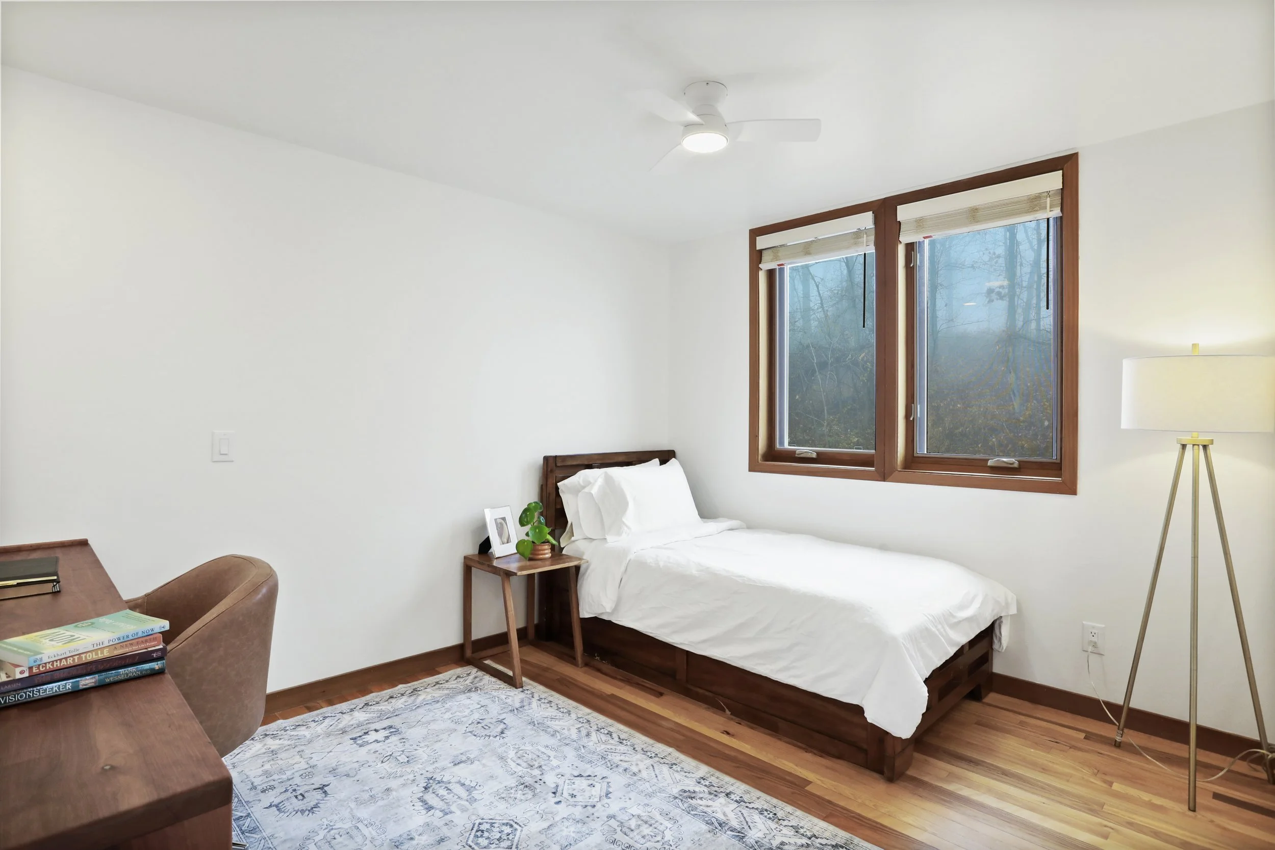 Minimalist bedroom with a single bed, wooden frame, white bedding, a small wooden nightstand with a plant and a picture, large windows with wood trim, a standing lamp, a ceiling fan, hardwood floors, and a light-colored area rug.