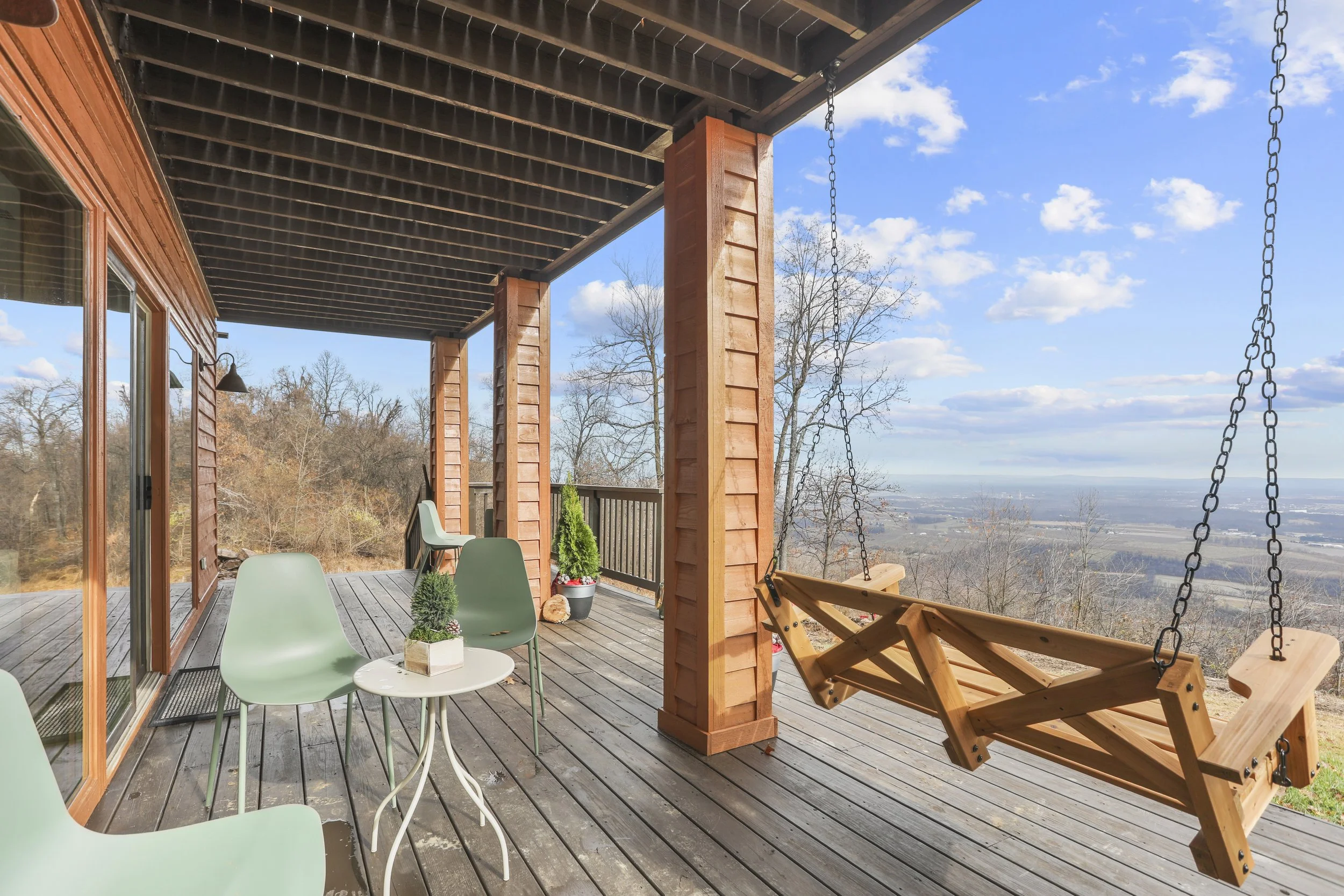 A wooden porch with a swing, chairs, and tables overlooking a scenic view of trees, a valley, and a partly cloudy sky.