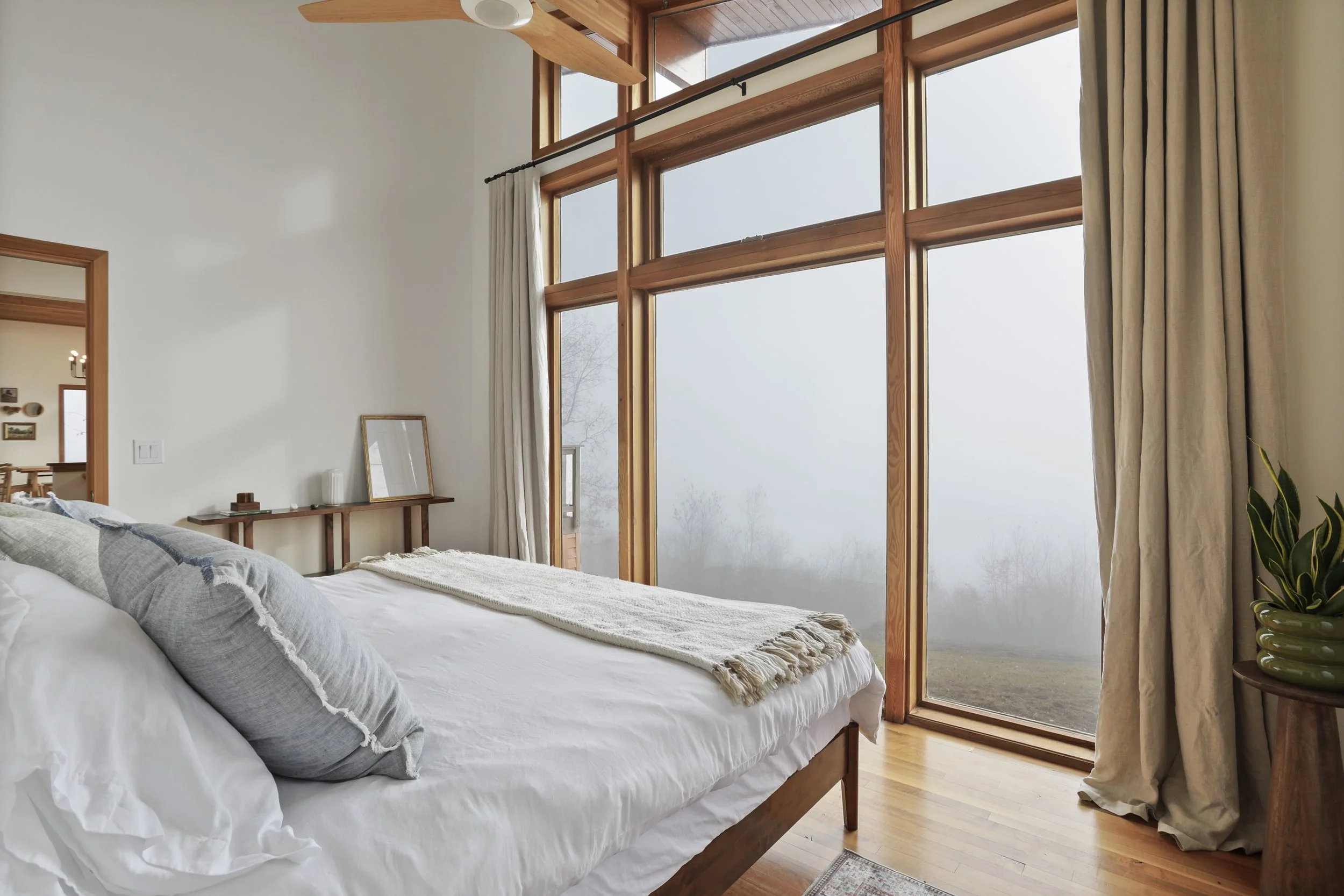 A cozy bedroom with large floor-to-ceiling windows showing foggy outdoor scenery. The bed has white linens and gray pillows. There is a wooden side table with a plant in a green pot and a mirror or picture frame leaning against the wall.