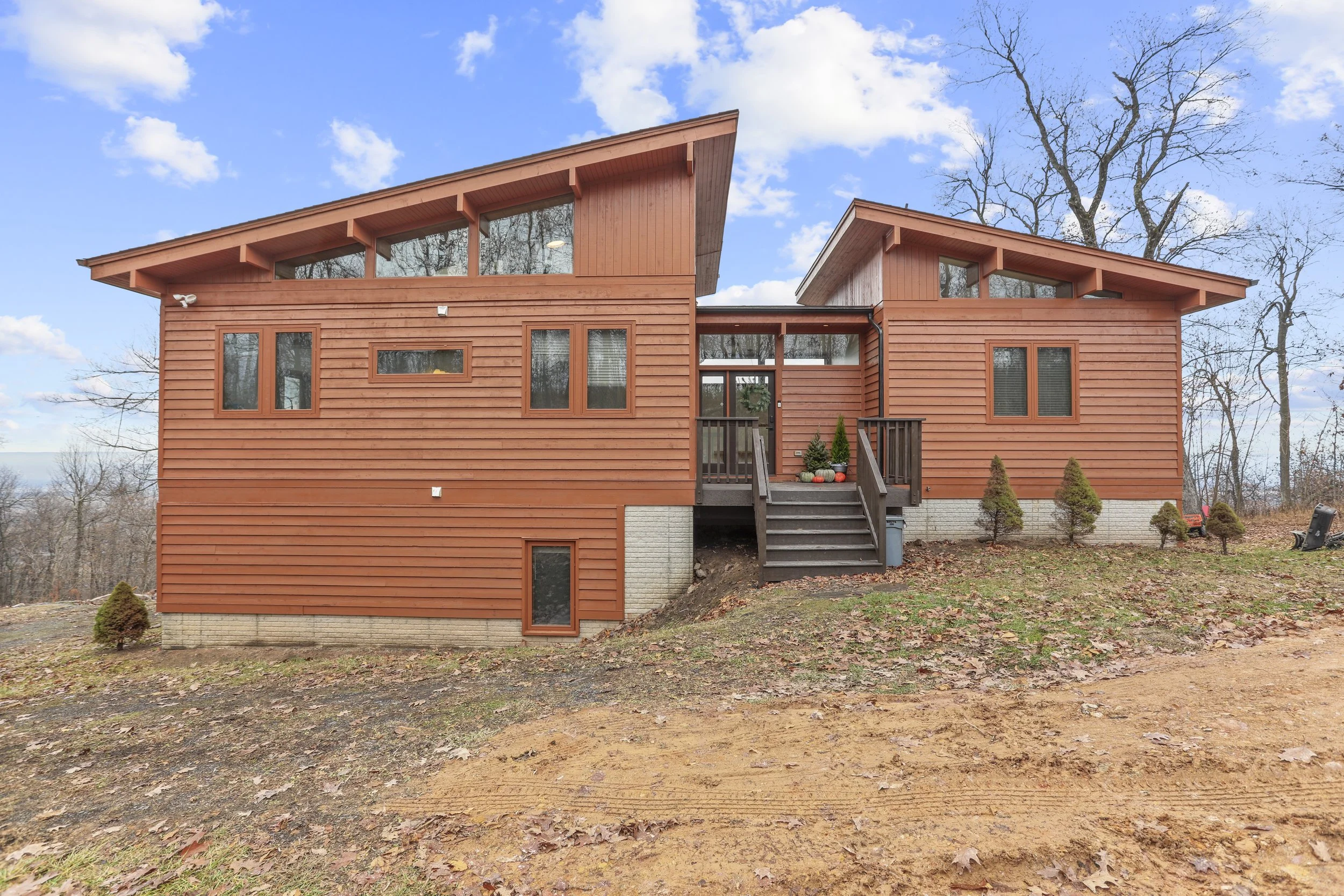 A modern two-story wooden house with a sloped roof, glass windows, and a small front porch with stairs, surrounded by a yard with sparse grass and small bushes, under a partly cloudy sky.