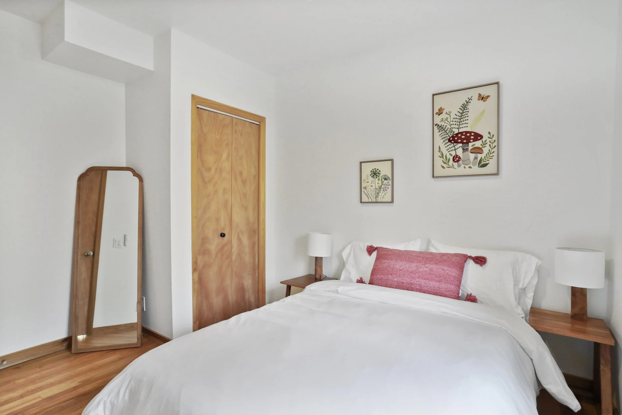 A bedroom with white walls, a wooden closet door, and a bed with white bedding and pink pillow. There are two small bedside tables with white lamps, a full-length mirror, and botanical art prints on the wall.