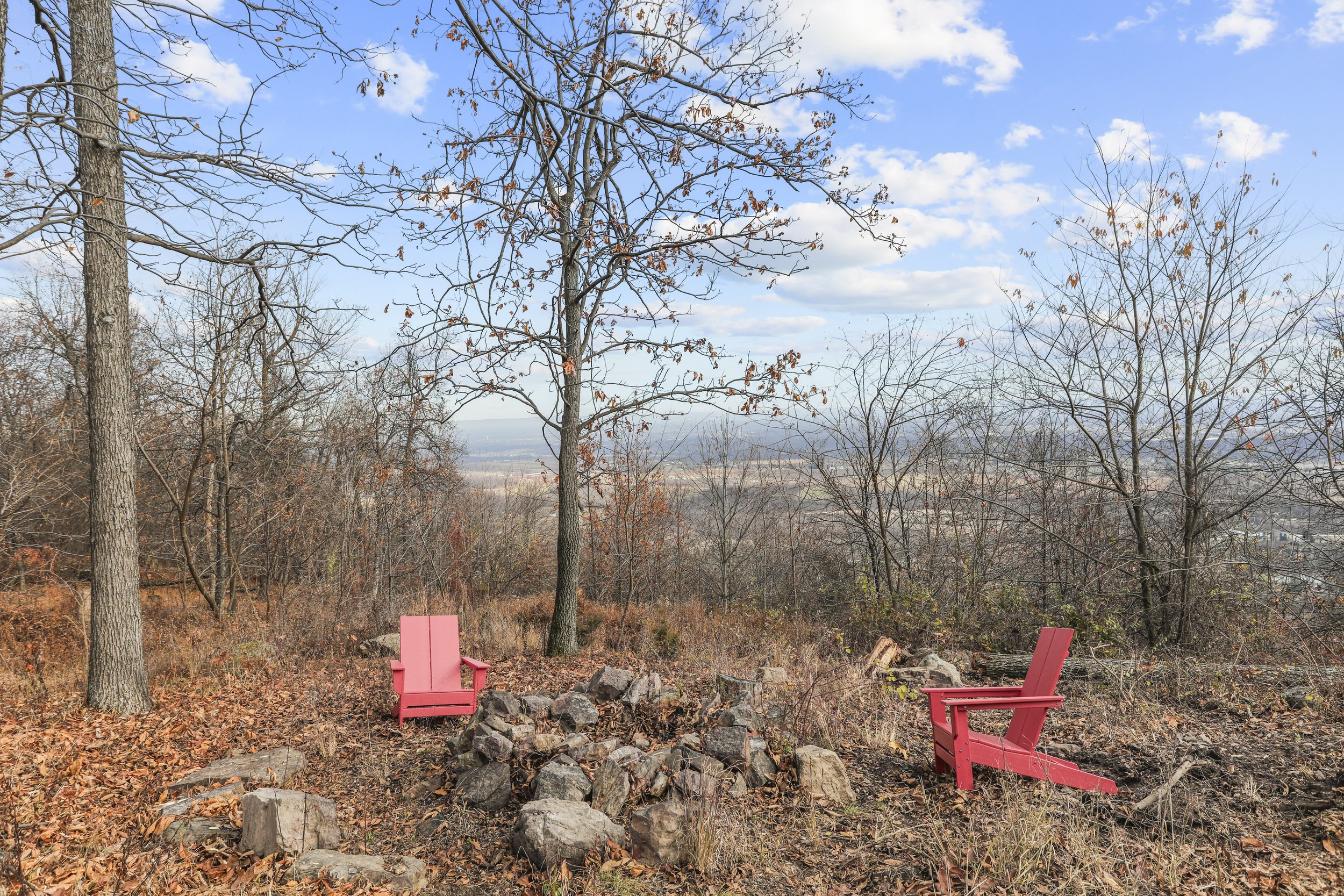 Two pink Adirondack chairs on a hillside surrounded by trees with brown leaves, with a stone fire pit between them and a view of the distant landscape under a partly cloudy sky.