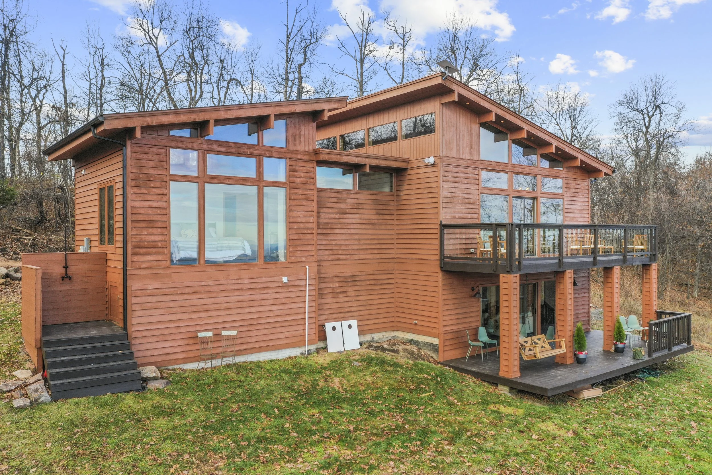 A modern, multi-level wooden house with large windows and two balconies, set in a wooded area with leafless trees and a grassy yard.