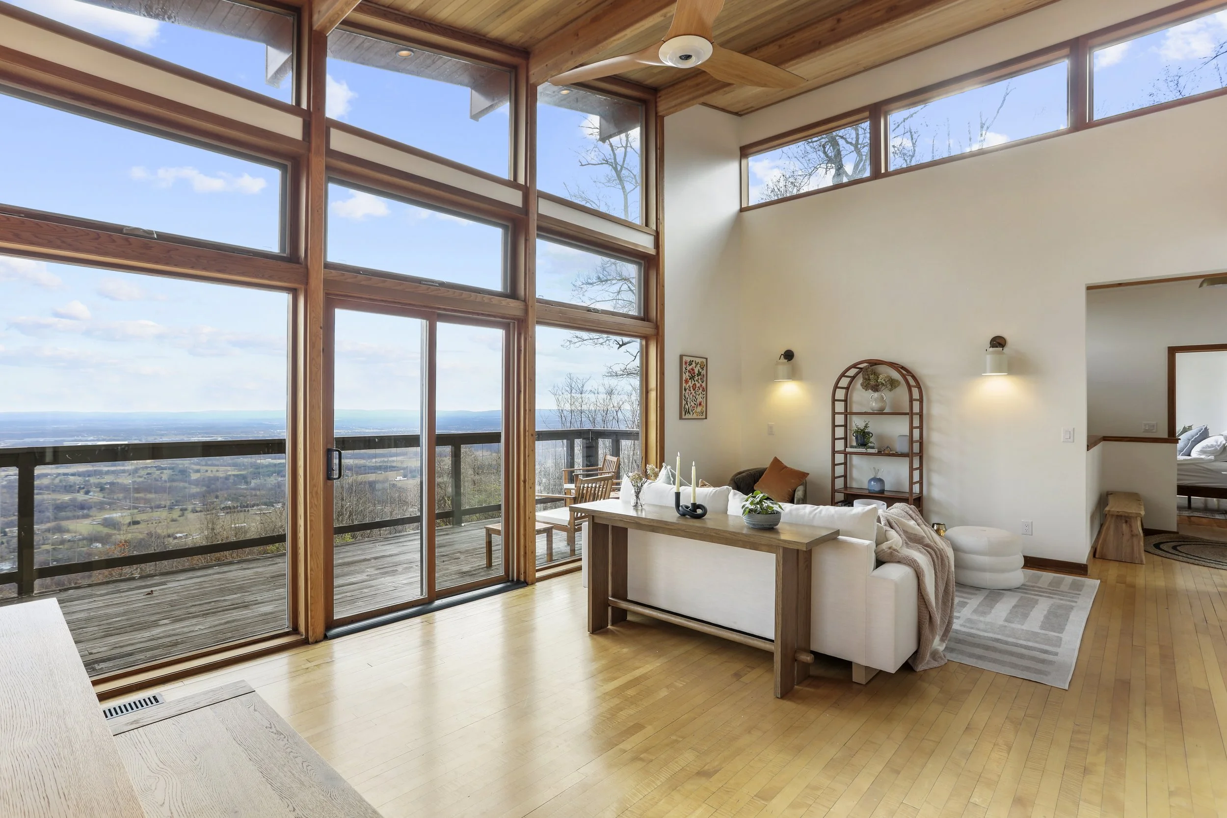 Living room with large glass windows and sliding door opening to a balcony with a scenic view. The room has light wooden floors, a white sofa, a wooden coffee table, and a decorative shelving unit. There are blue and white accents, and a ceiling fan 