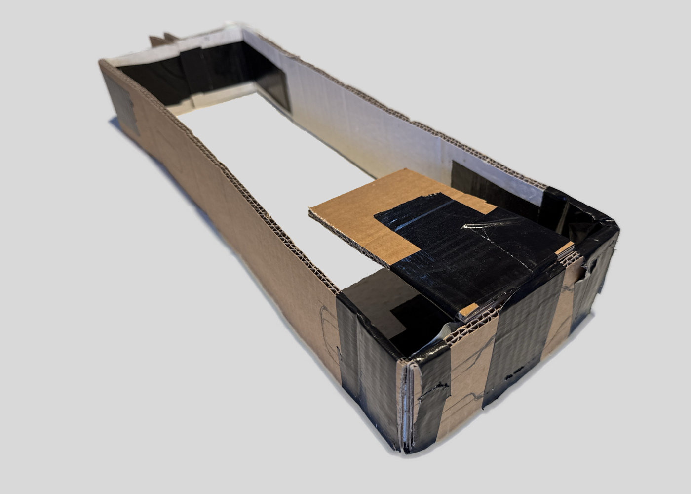 Empty cardboard box with black tape on edges and a piece of brown cardboard inside on a plain gray background. This is the second step of the procedure, a more advanced model in cardboard.
