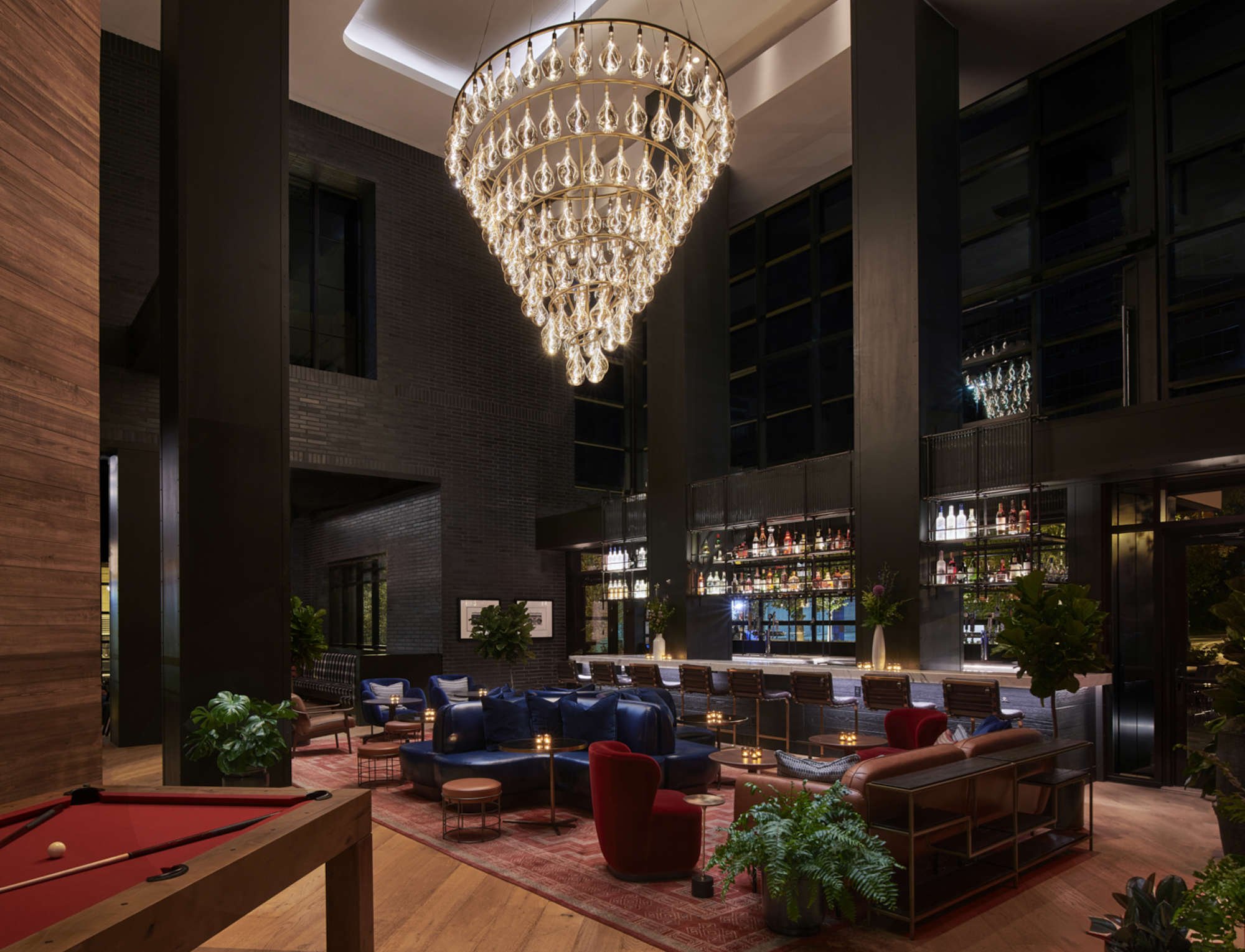 A luxurious hotel lobby with a large crystal chandelier, a bar area with colorful bottles, and comfortable seating including blue, red, and beige chairs, surrounded by green plants and warm lighting.