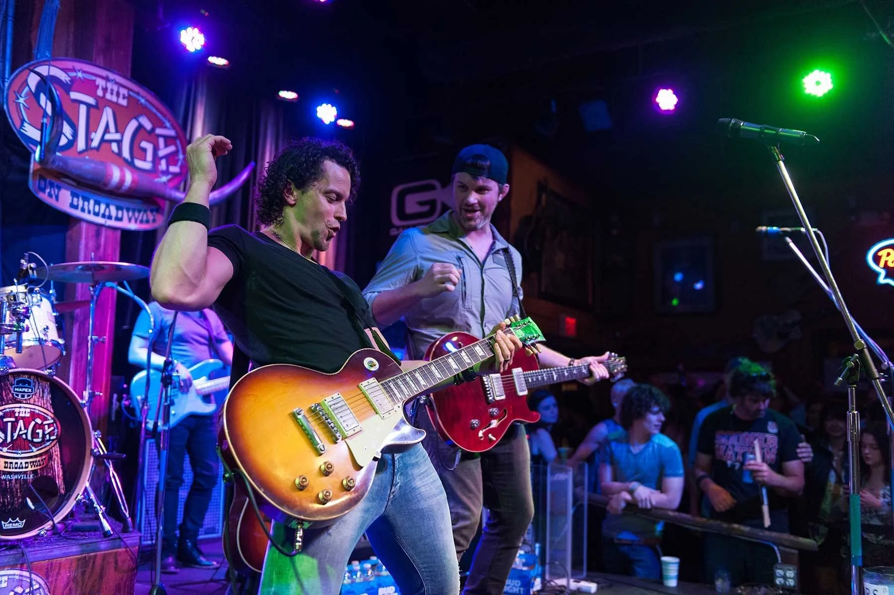 Musicians playing guitars on stage at a live music venue with audience watching.