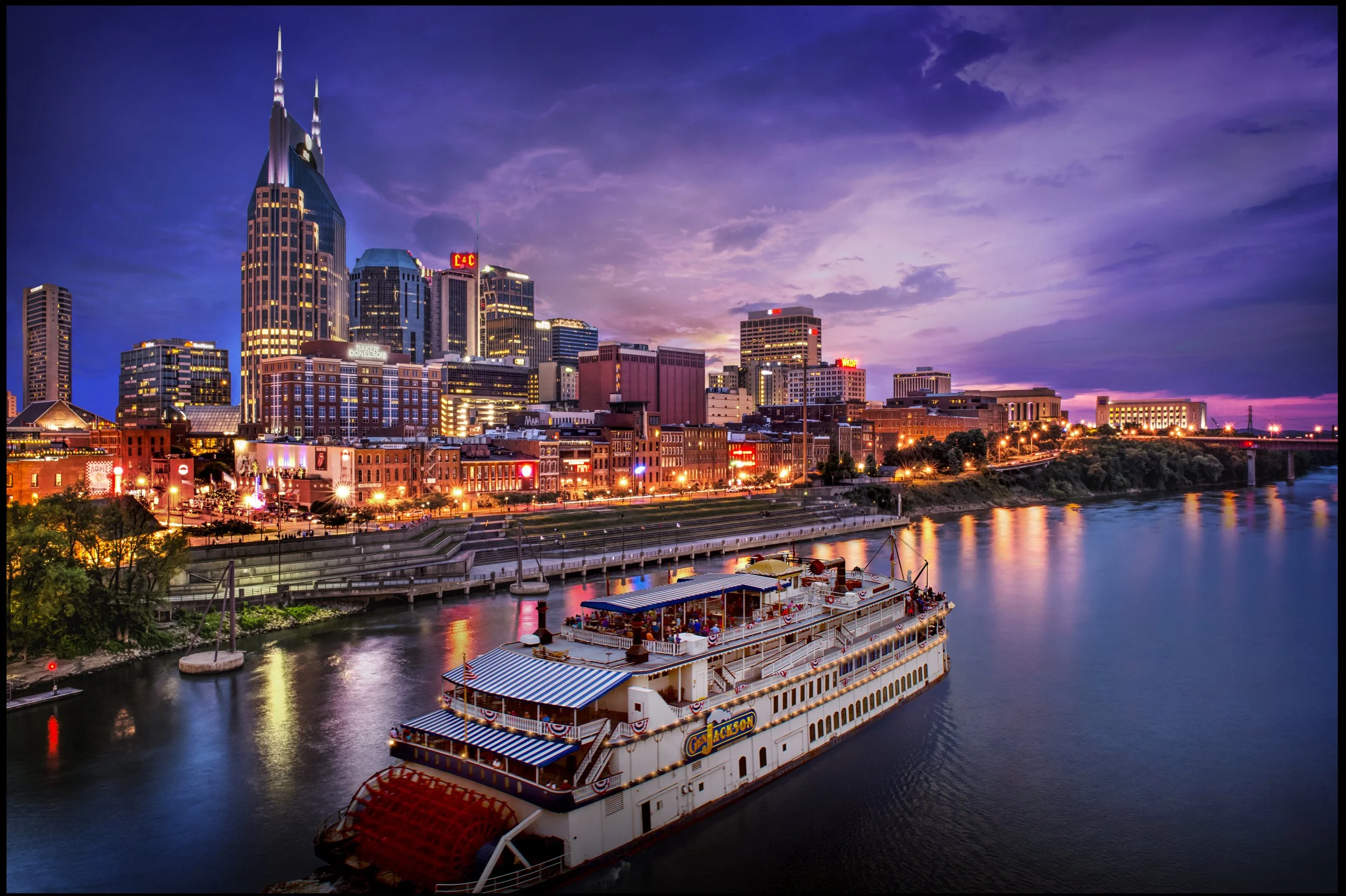 City skyline at sunset with tall buildings and a boat on the river in Nashville, Tennessee.