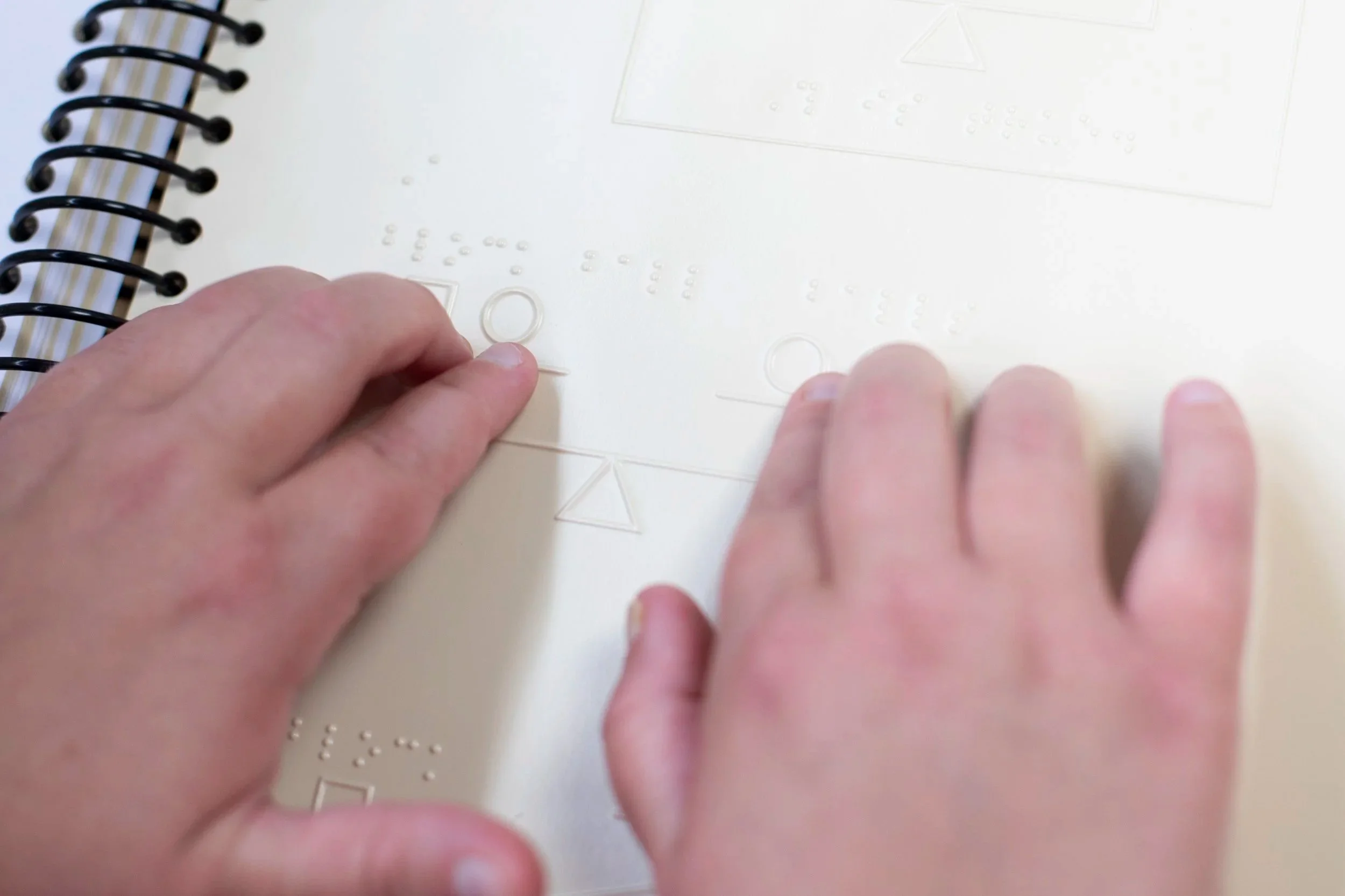 Close-up of hands handling clear braille embossing labels on a white surface, with a spiral-bound notebook in the background.