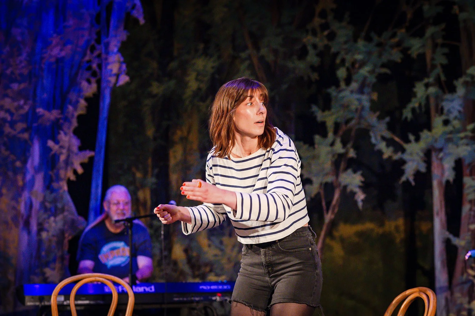 A woman with shoulder-length brown hair in a striped shirt and black shorts dancing on stage with a man playing a keyboard in the background surrounded by trees.