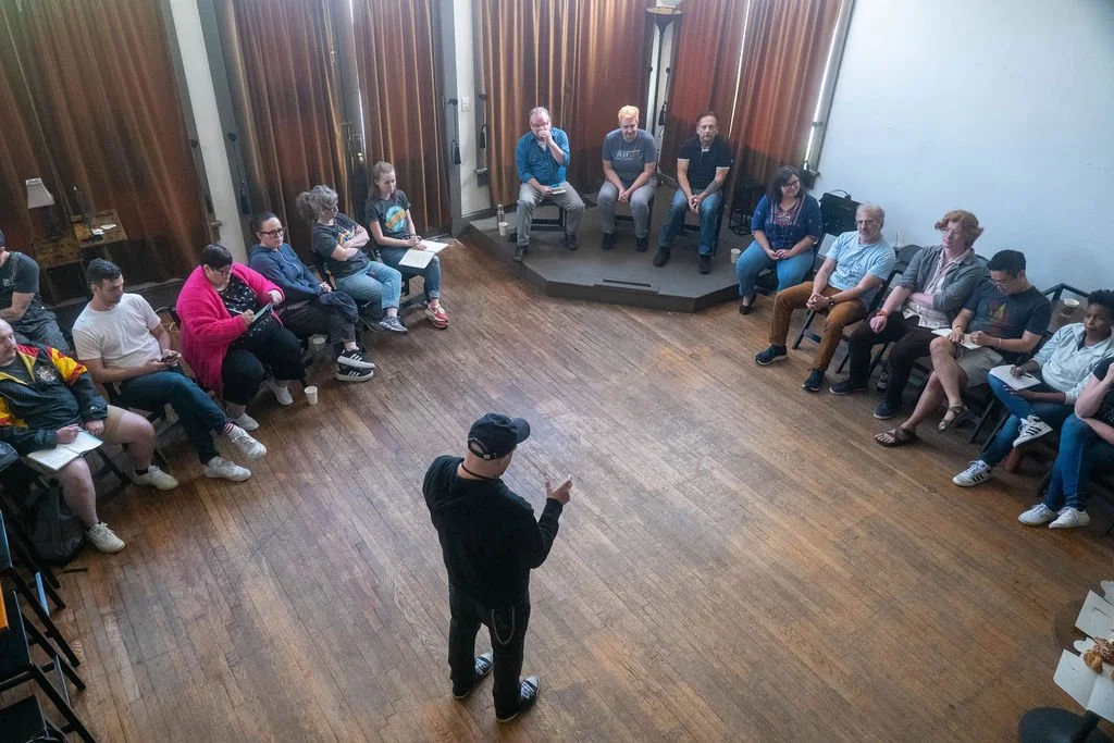 A man standing in the center of a circular room, speaking to a group of people seated along the walls.