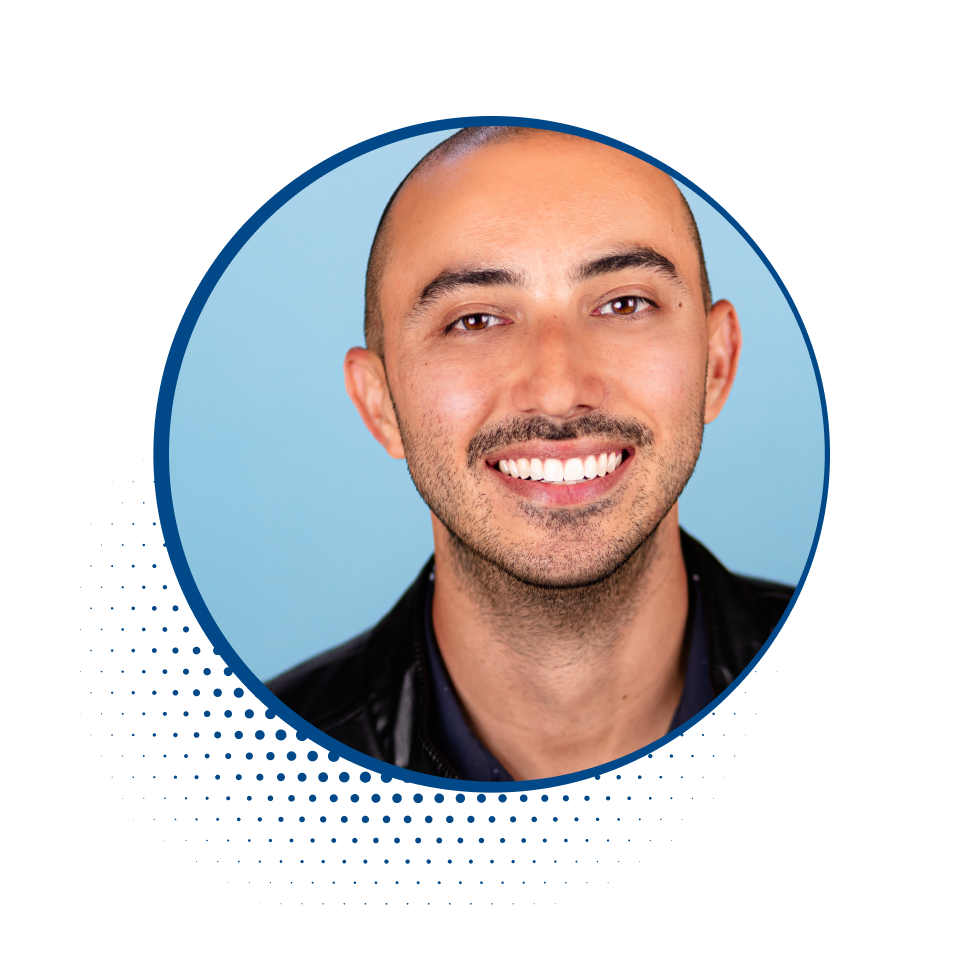 Portrait of a smiling man with short dark hair, light skin, and a stubble beard, wearing a black jacket against a blue background.